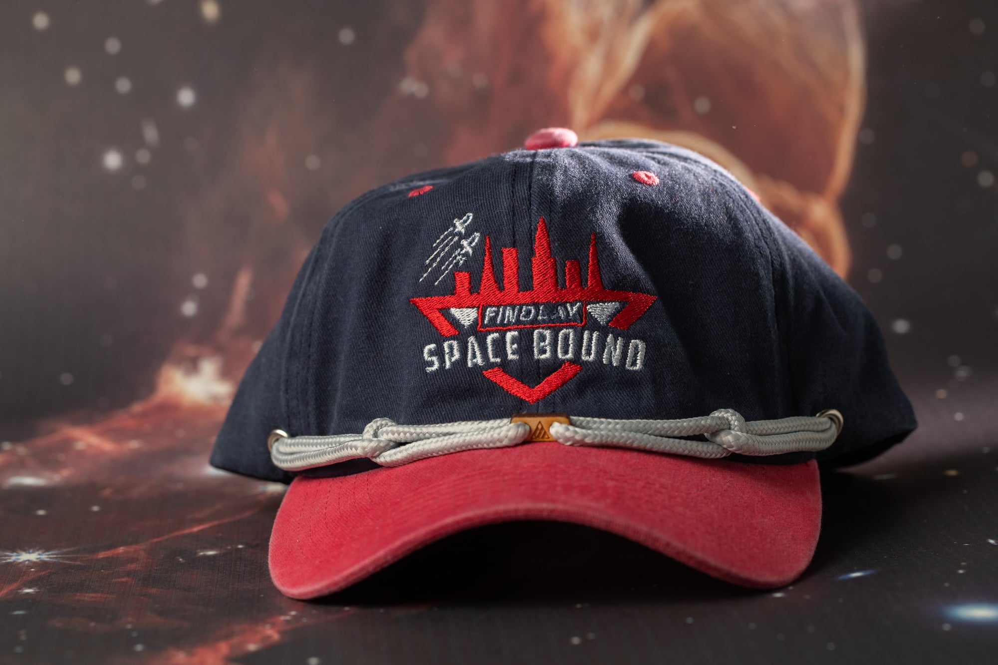 A navy blue baseball cap with a red brim and a logo that says "FINDlay Space Bound" on it, placed on a dark background with a starry sky.