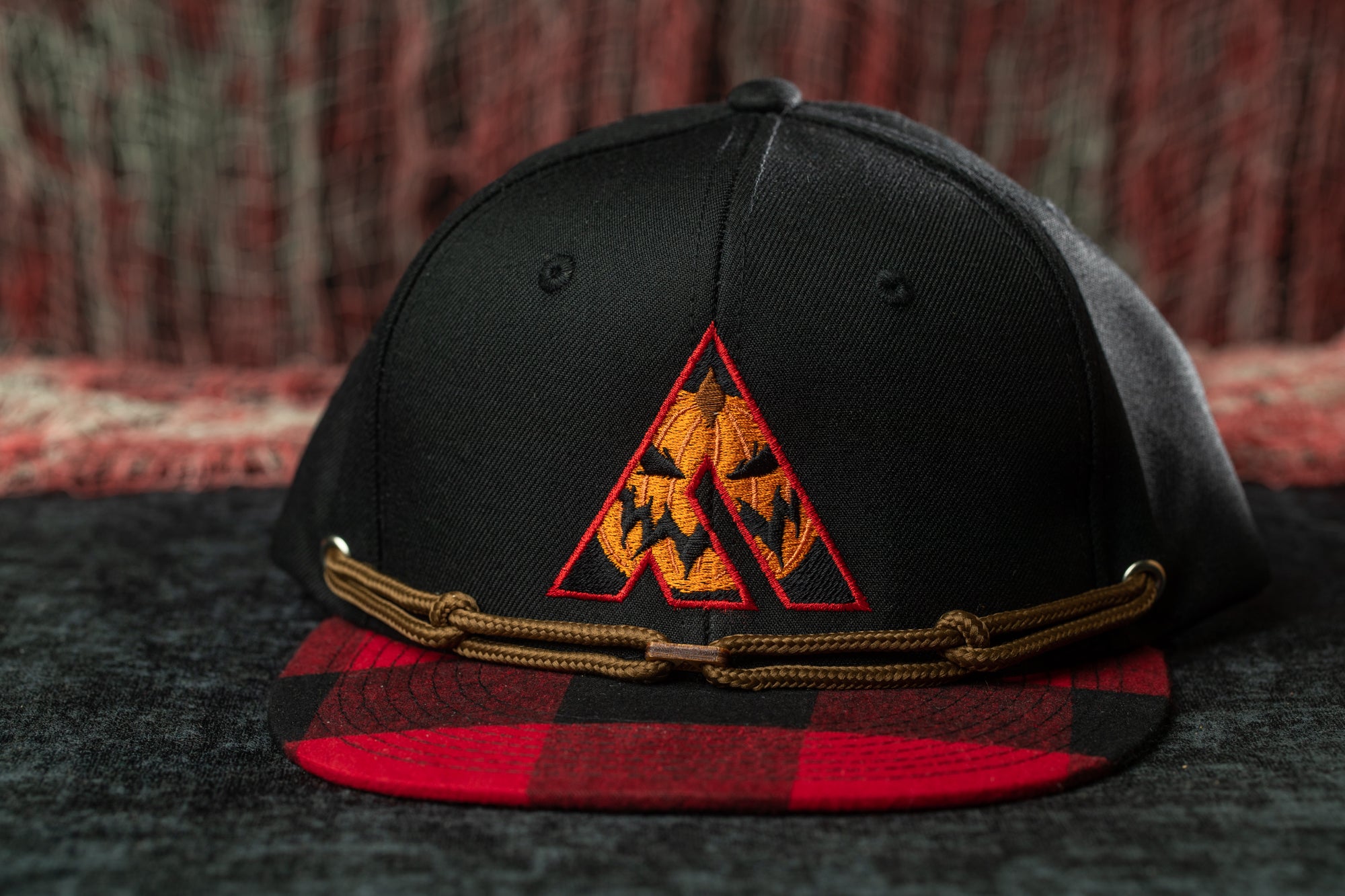 A black baseball cap with a red and black plaid pattern and a pumpkin-themed logo on the front.