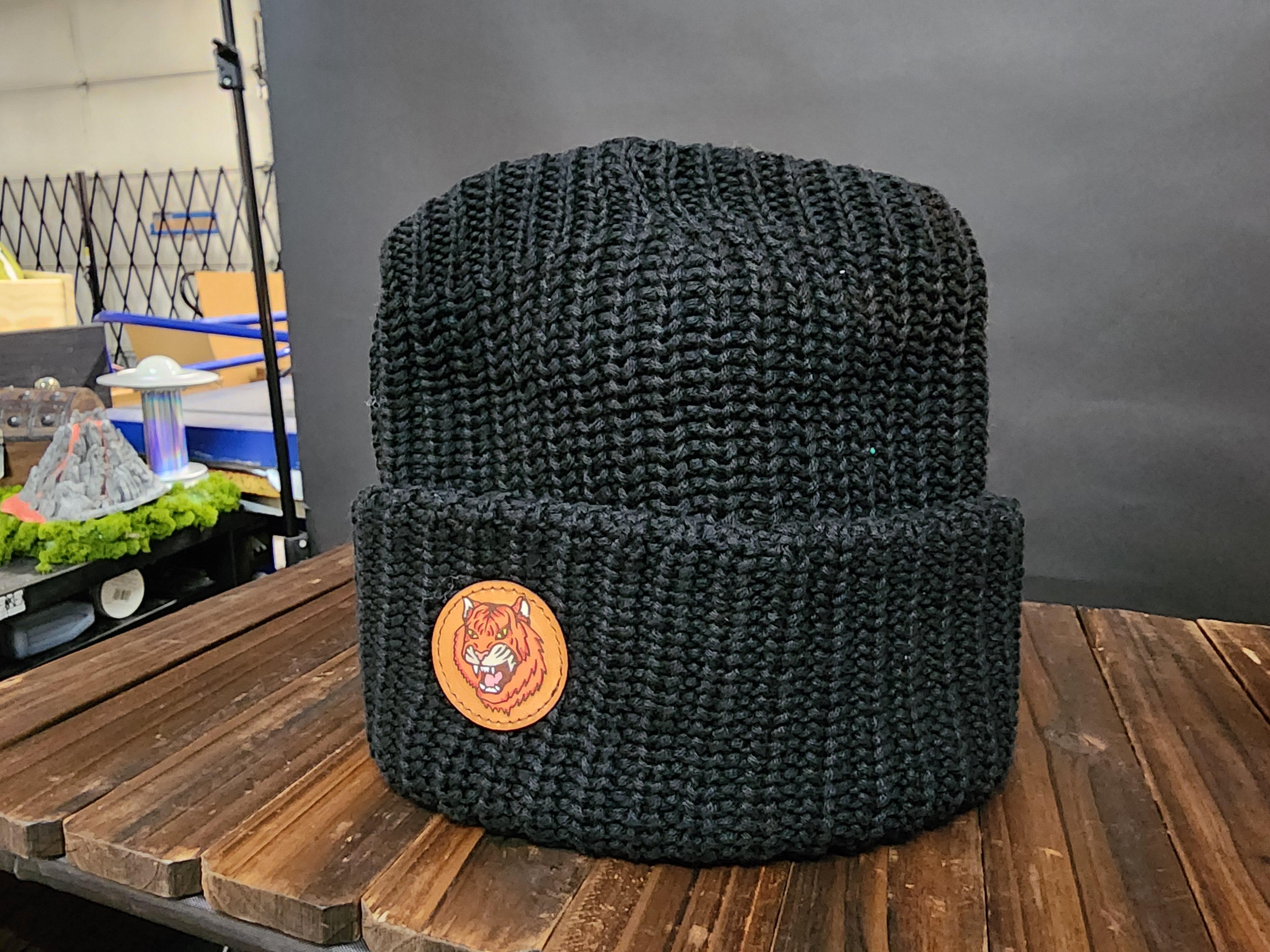A black knit beanie with a tiger logo is displayed on a wooden surface.