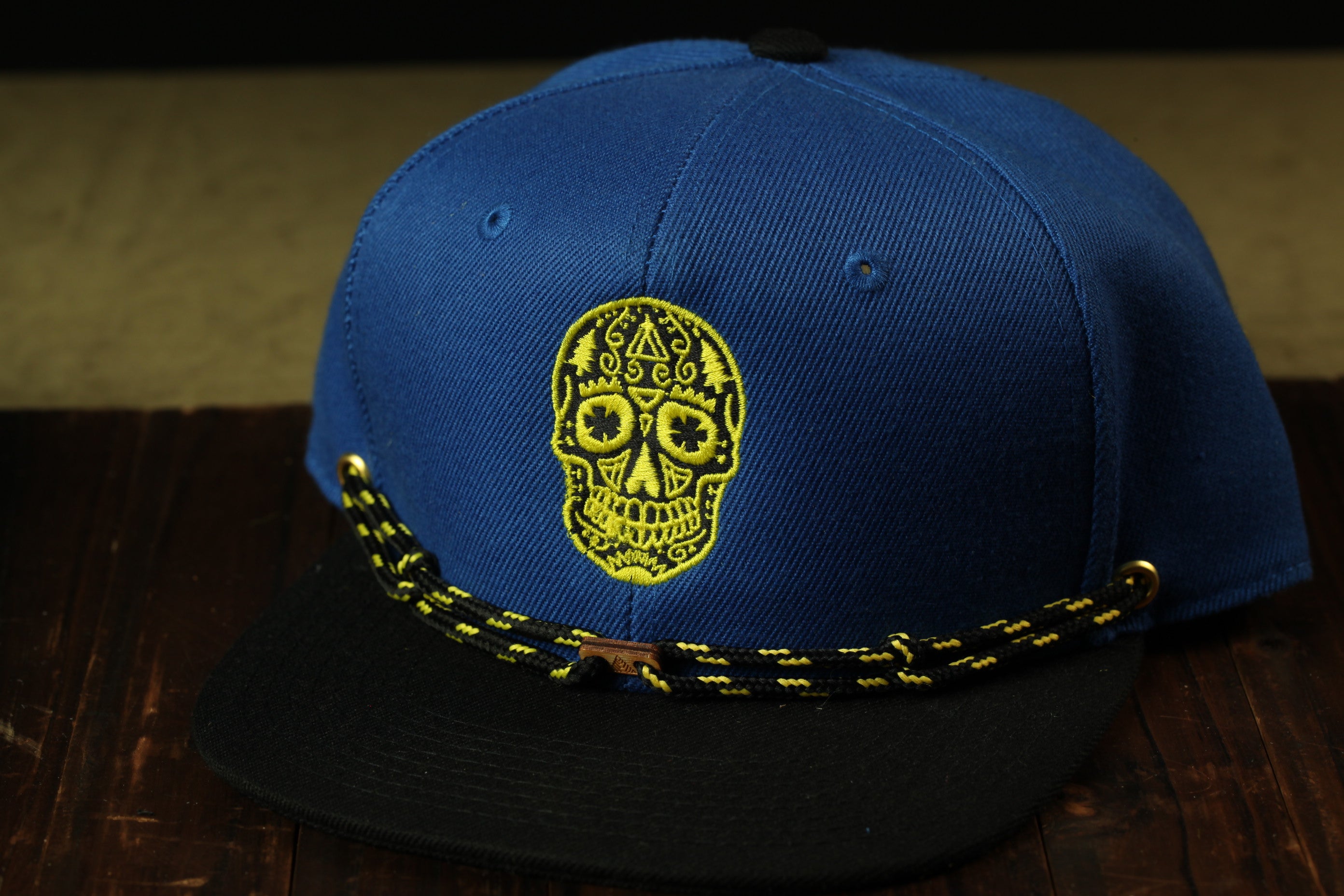 A blue baseball cap with a yellow skull embroidered on the front, sitting on a wooden surface.