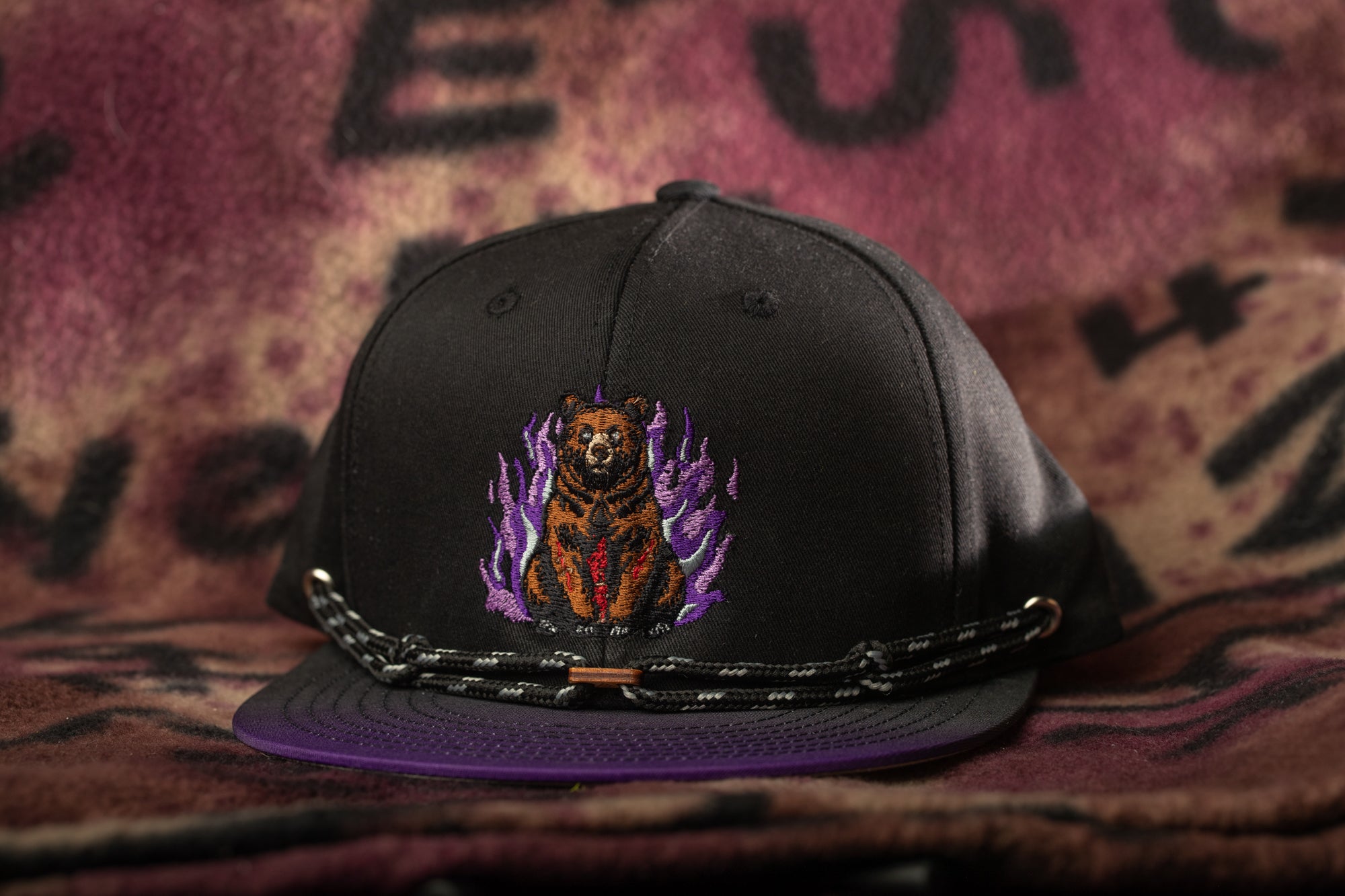 A black baseball cap with a purple and orange embroidered design on the front, featuring a bear sitting amidst flames.