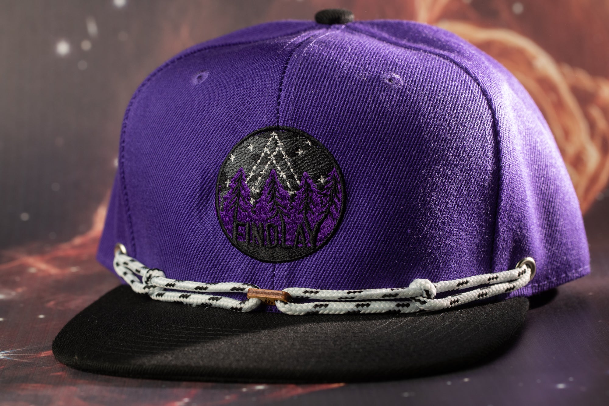 A purple baseball cap with a black brim and a white rope strap, featuring a circular logo with a mountain and trees design.