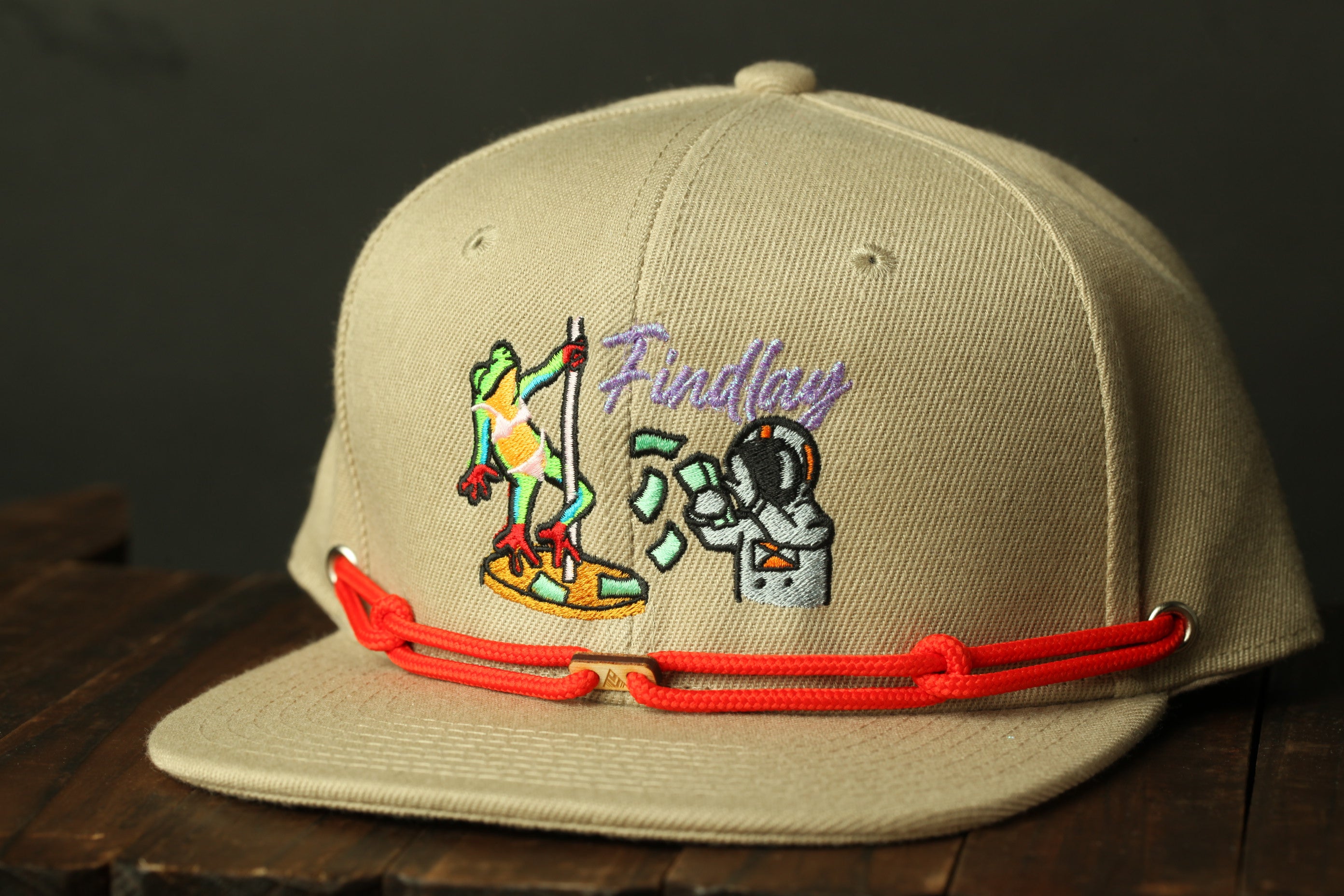 A beige baseball cap with a red strap and a colorful embroidered design featuring a frog and a person in a spacesuit.