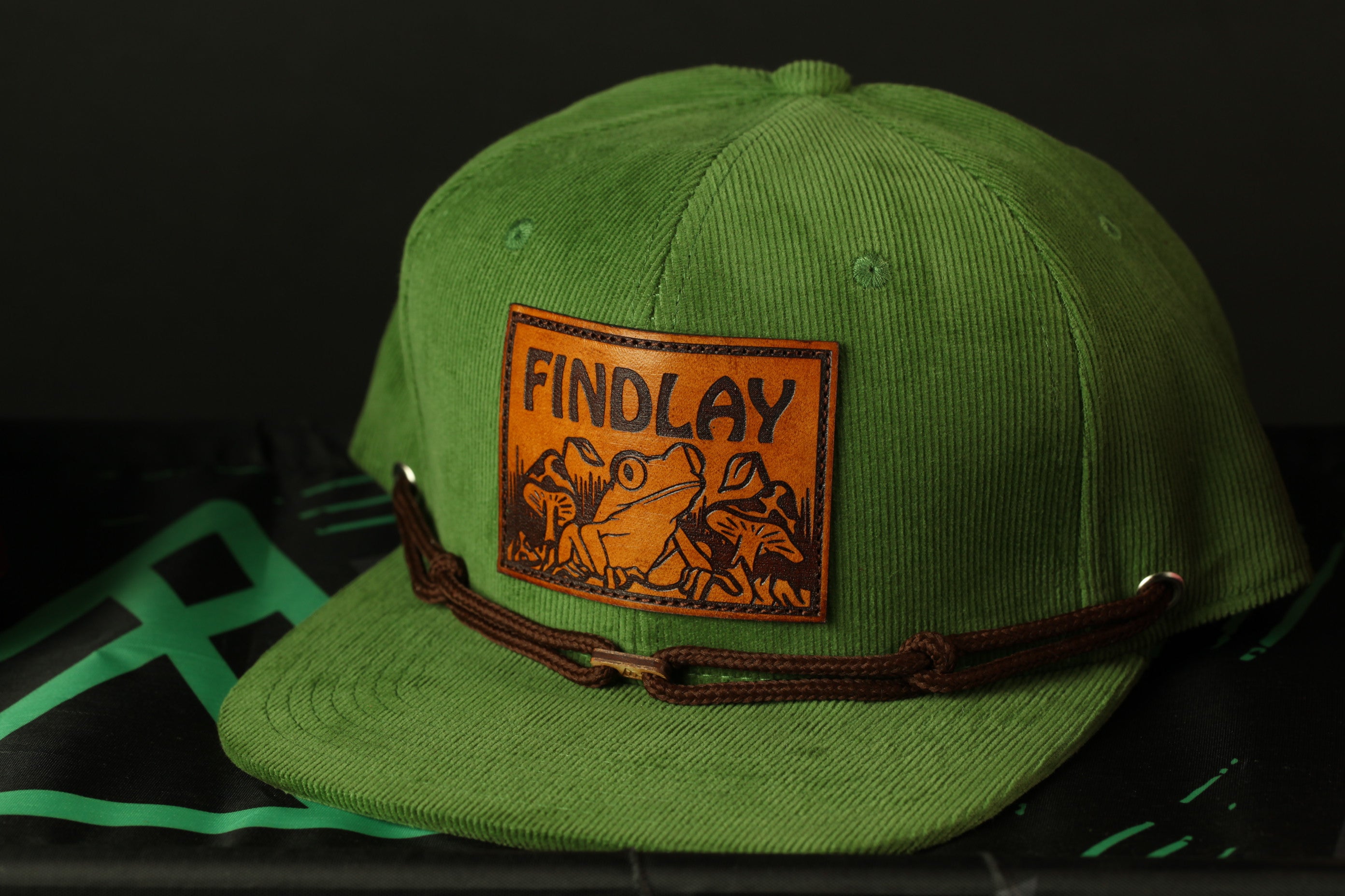 A green baseball cap with a brown leather patch on the front, displaying the text "FINDLAY" and an illustration of a frog.
