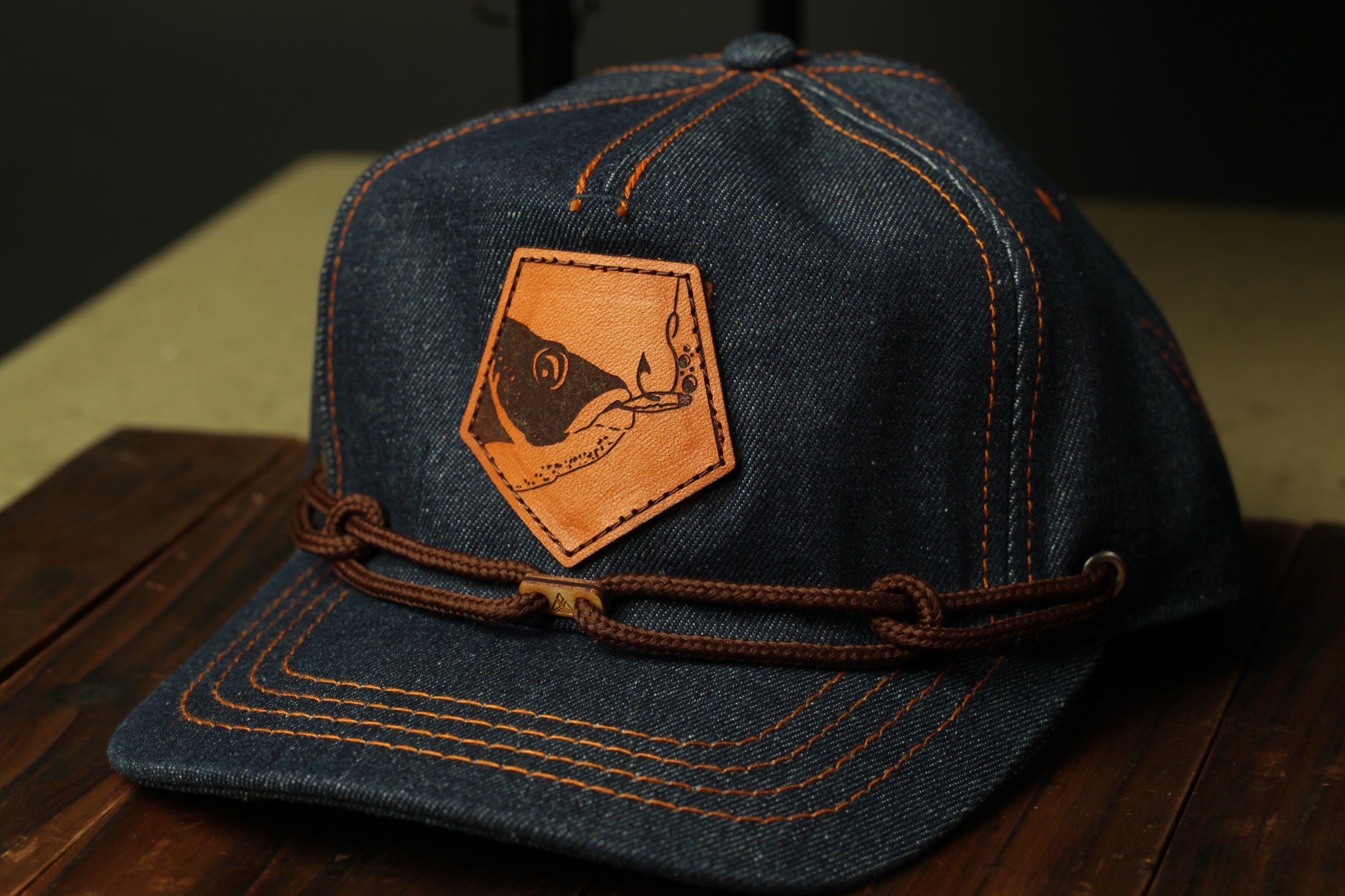 A denim baseball cap with a leather patch featuring a fish design, placed on a wooden surface.