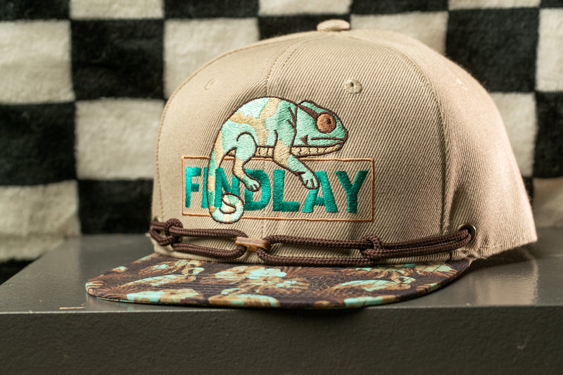 A beige baseball cap with a chameleon design and the word "FINDlay" printed on it, sitting on a surface with a checkered pattern in the background.