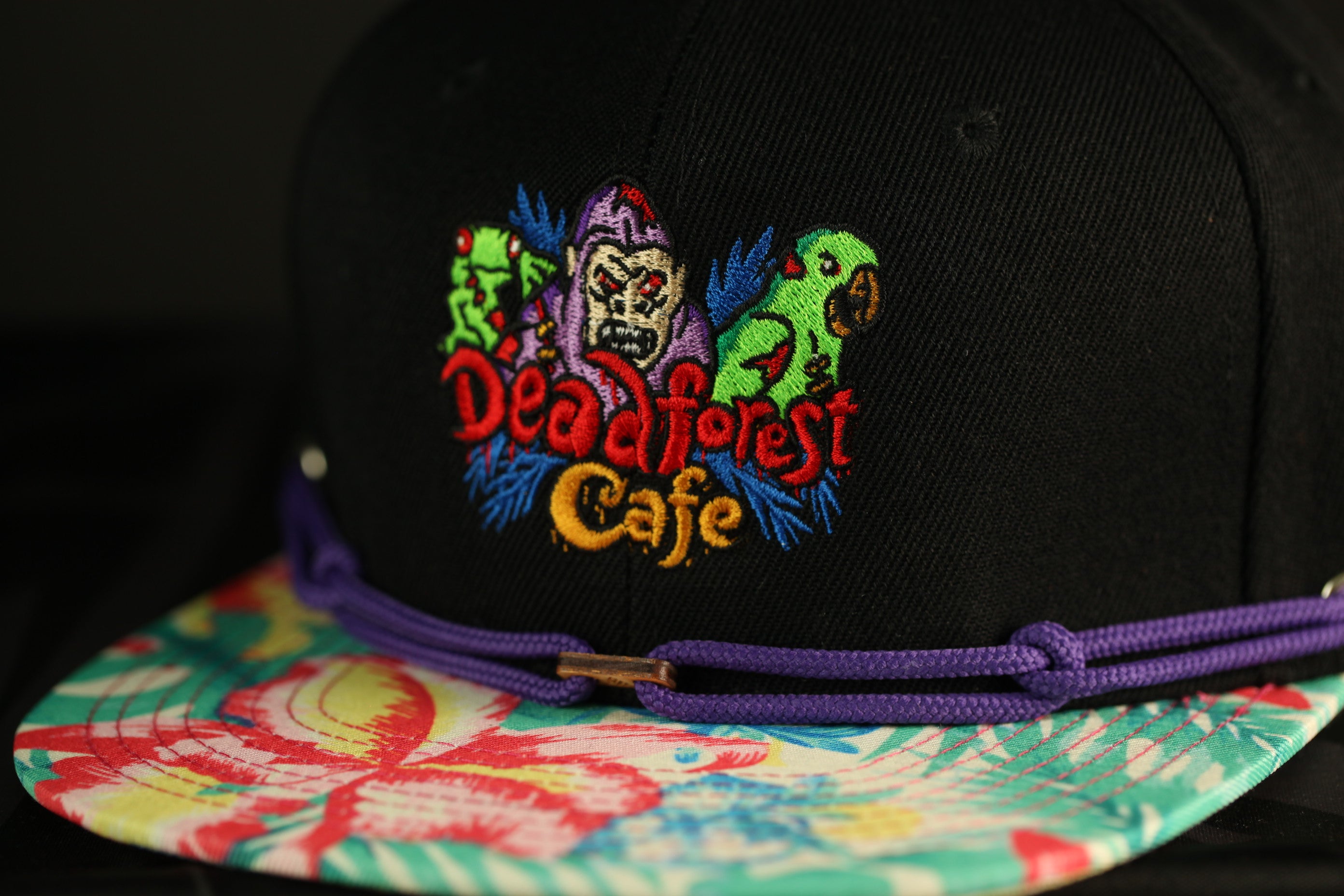 A close-up of a black baseball cap with a colorful floral pattern and a logo for "Dead Forest Cafe" embroidered on it.