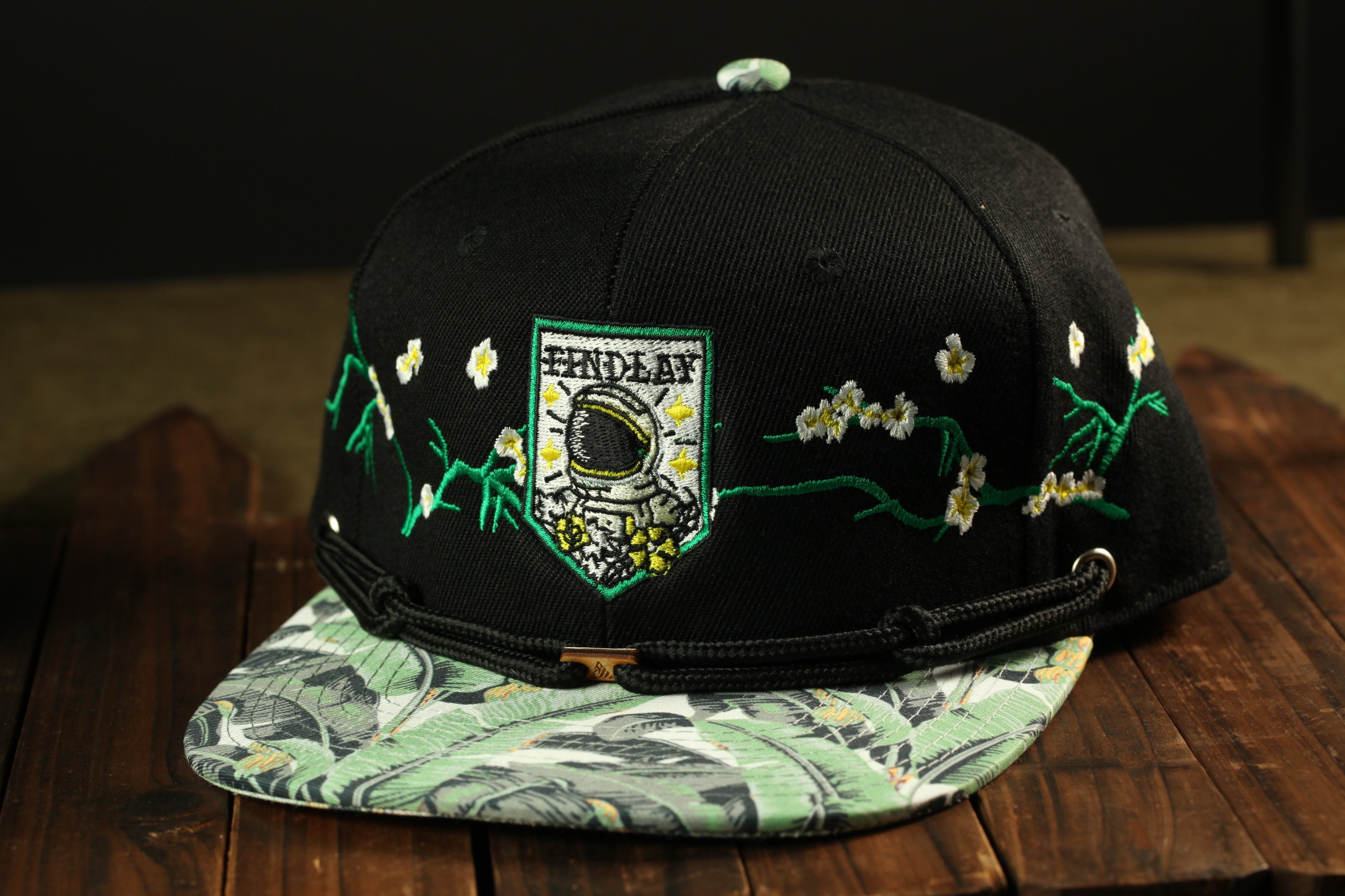A black baseball cap with a floral pattern and a small embroidered patch on the front, placed on a wooden surface.