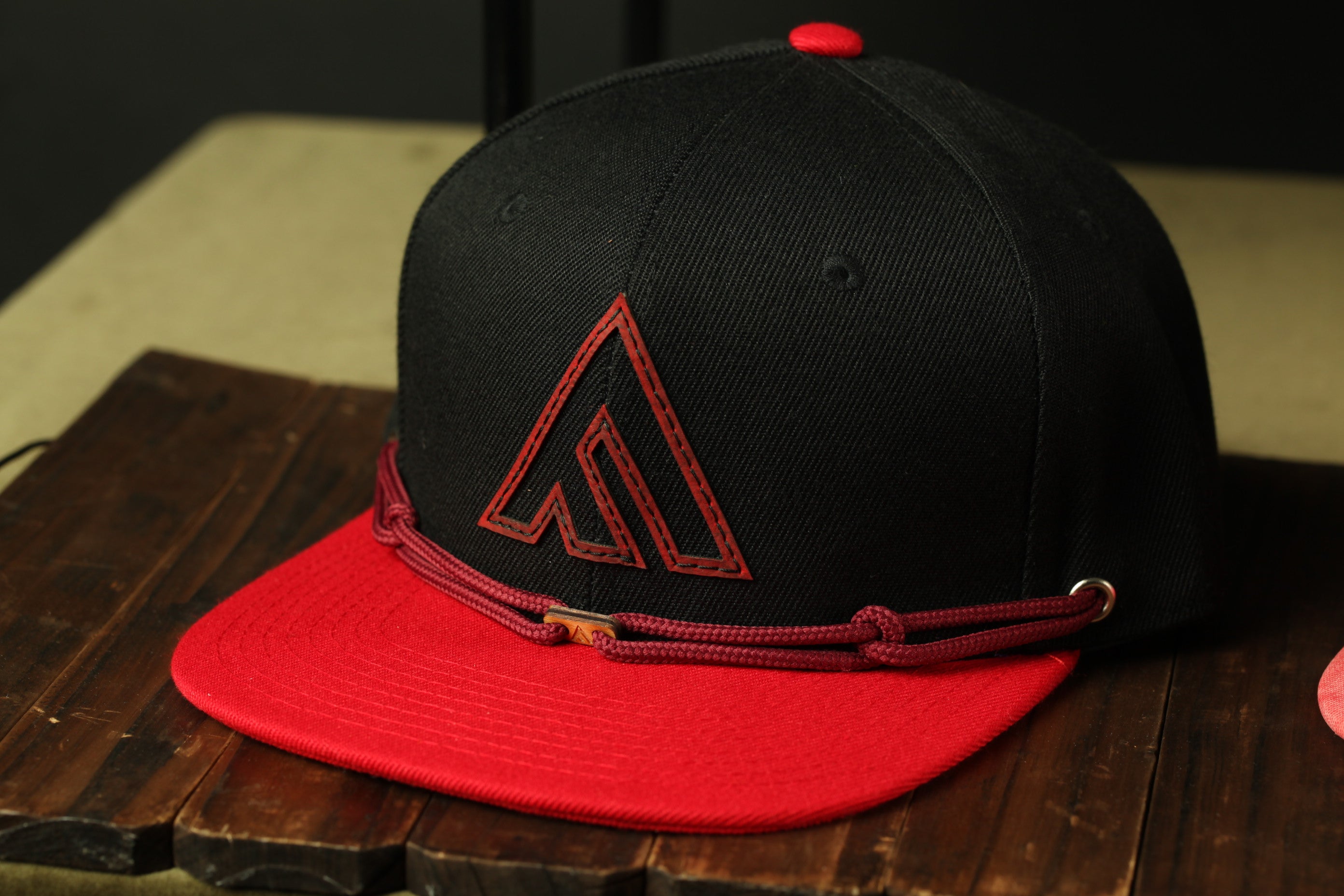 A black baseball cap with a red brim and a red logo on the front, placed on a wooden surface.
