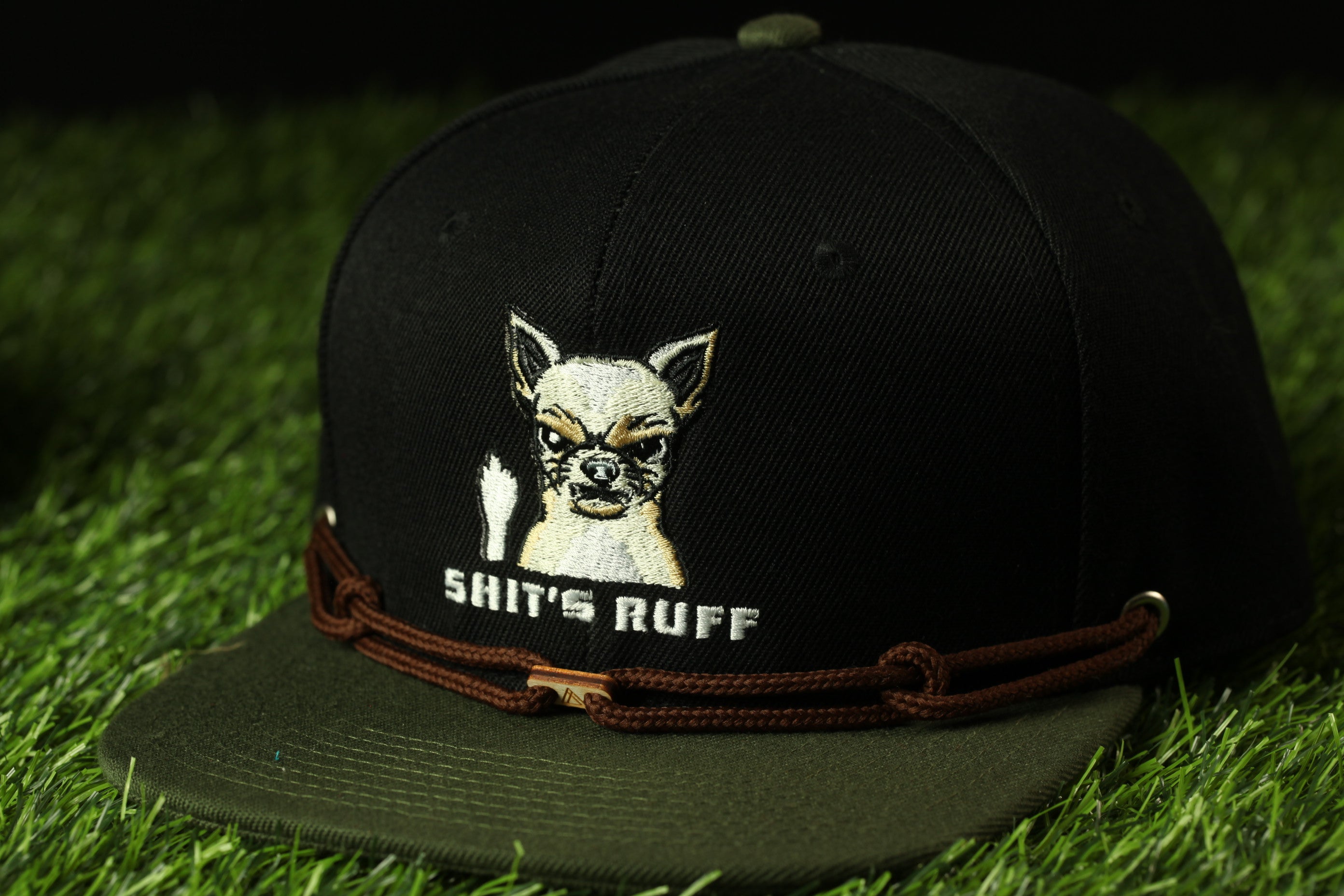 A black baseball cap with a graphic of a dog and the text "SHIT'S RUFF" printed on it, placed on a grassy surface.