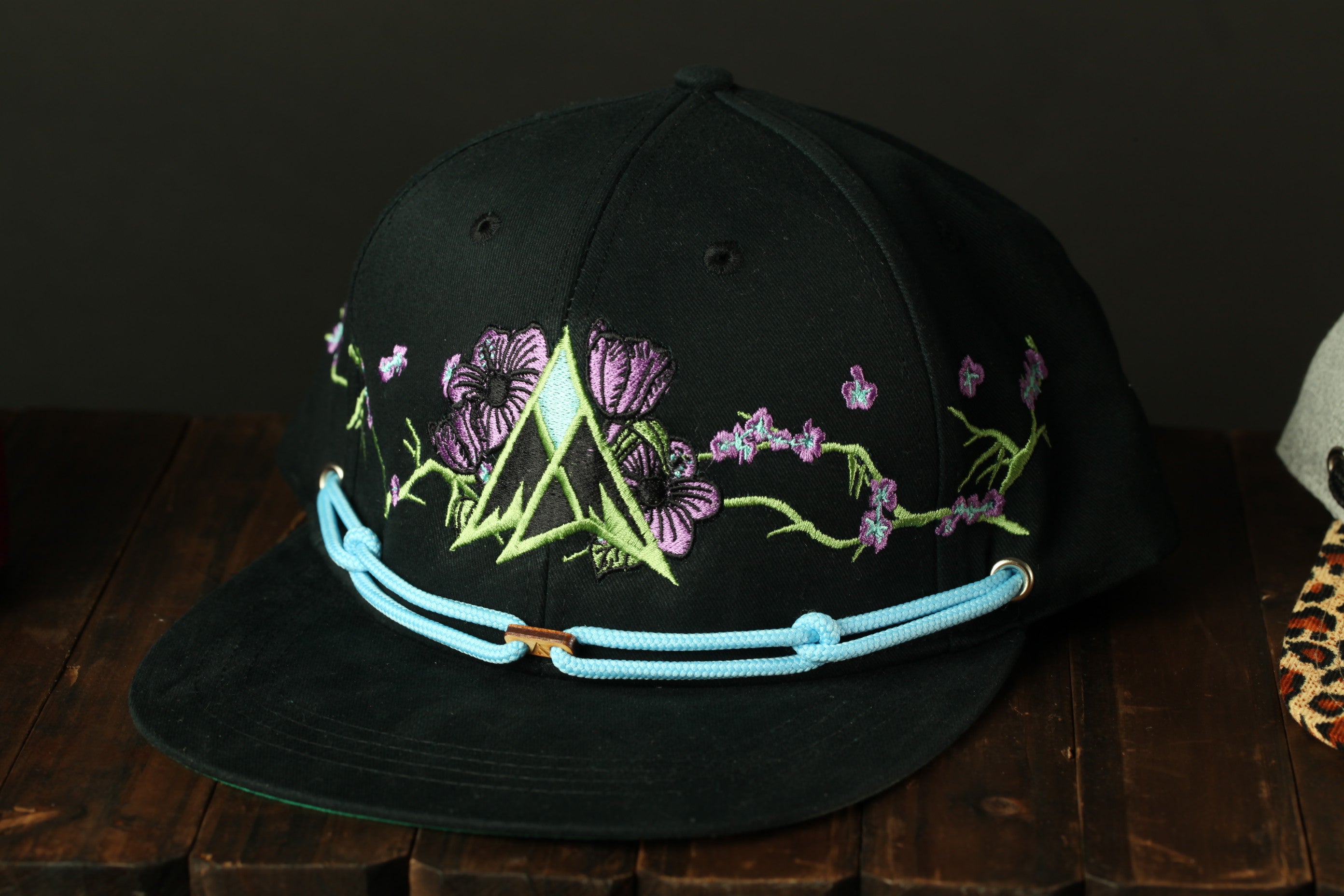 A black baseball cap with a floral embroidered design on the front, sitting on a wooden surface.