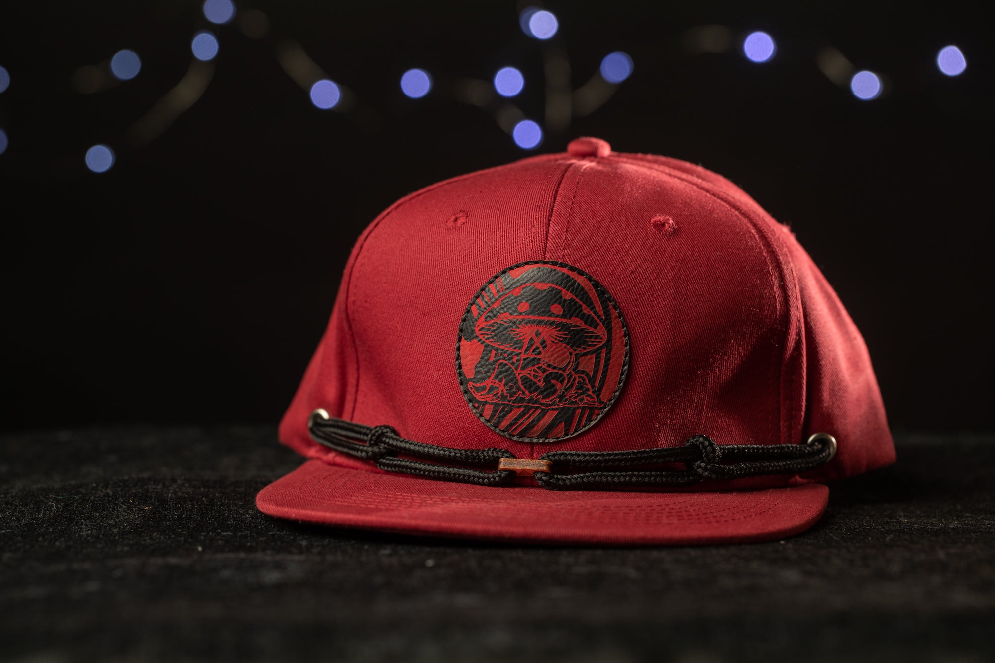 A red baseball cap with a black mushroom design on the front, placed on a black surface against a background of blue lights.