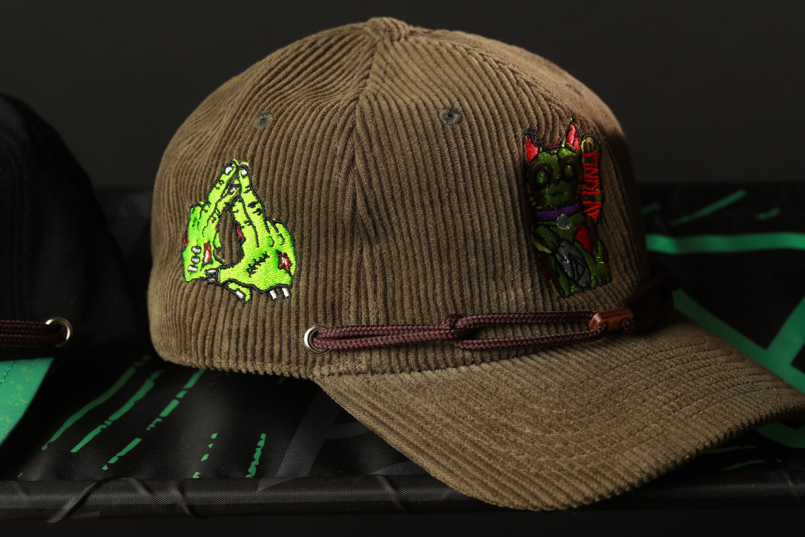 A brown baseball cap with various colorful and unique embroidered designs, including a green monster-like figure and a red and black character, is displayed on a surface.