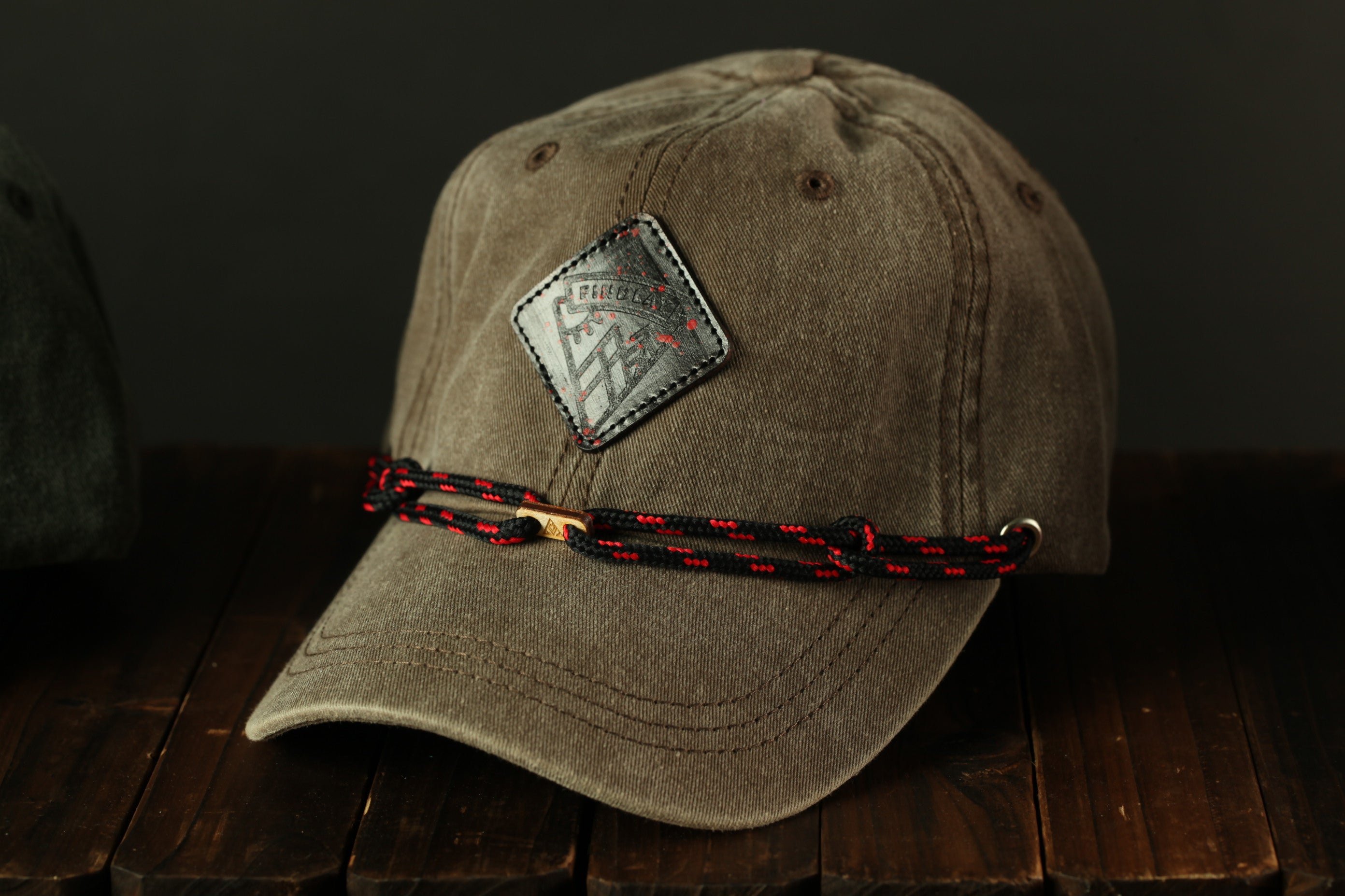 A brown baseball cap with a black and red braided strap is displayed on a wooden surface.