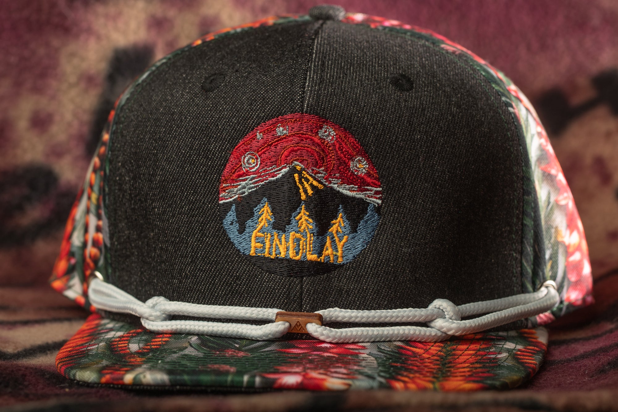 A black baseball cap with a colorful floral pattern and a red and yellow embroidered logo on the front.