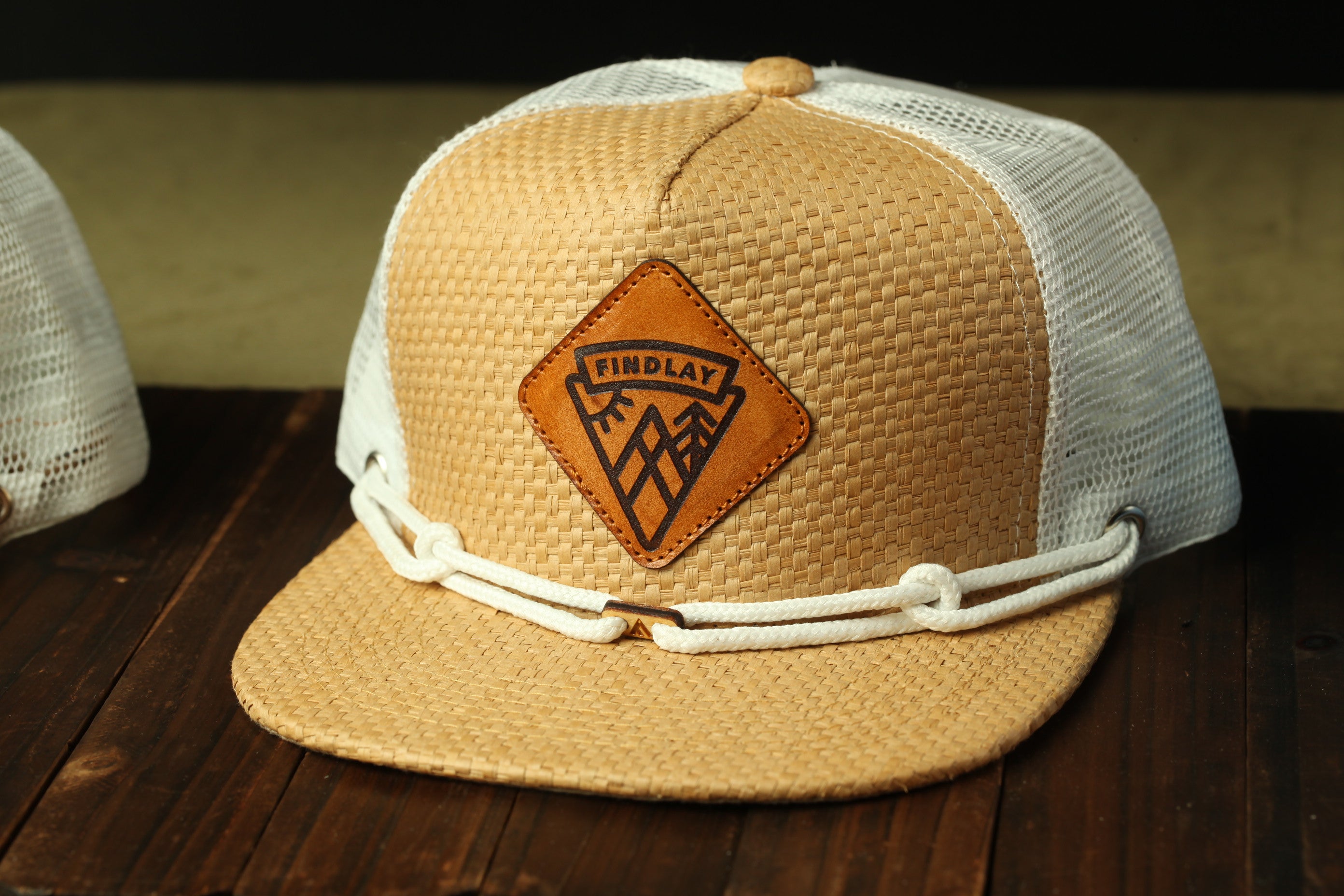 A tan baseball cap with a woven texture and a leather patch featuring a mountain design, placed on a wooden surface.