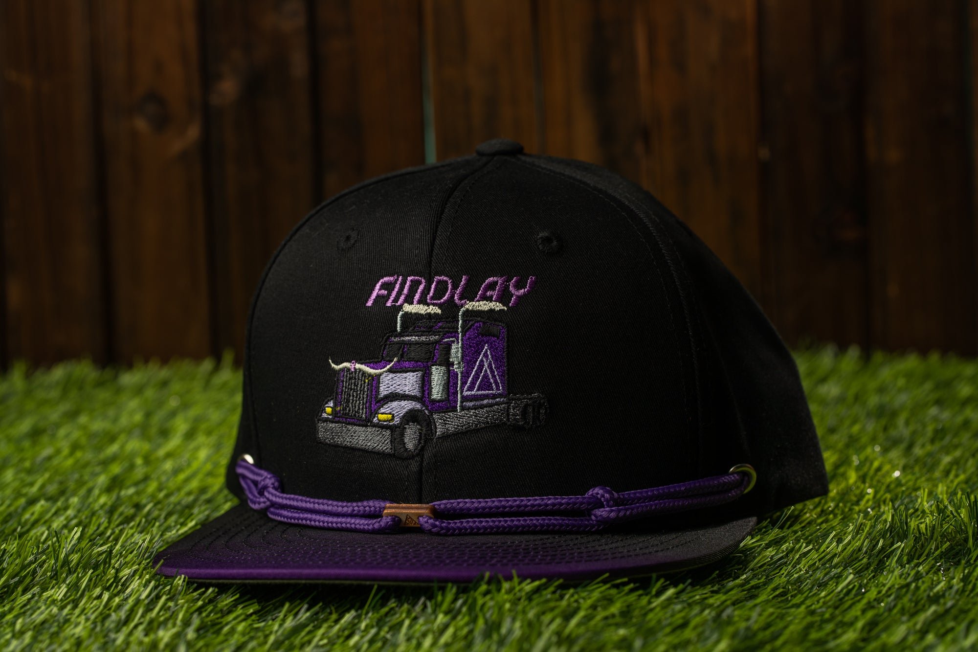 A black baseball cap with a purple and white truck design on it, placed on a grassy surface.