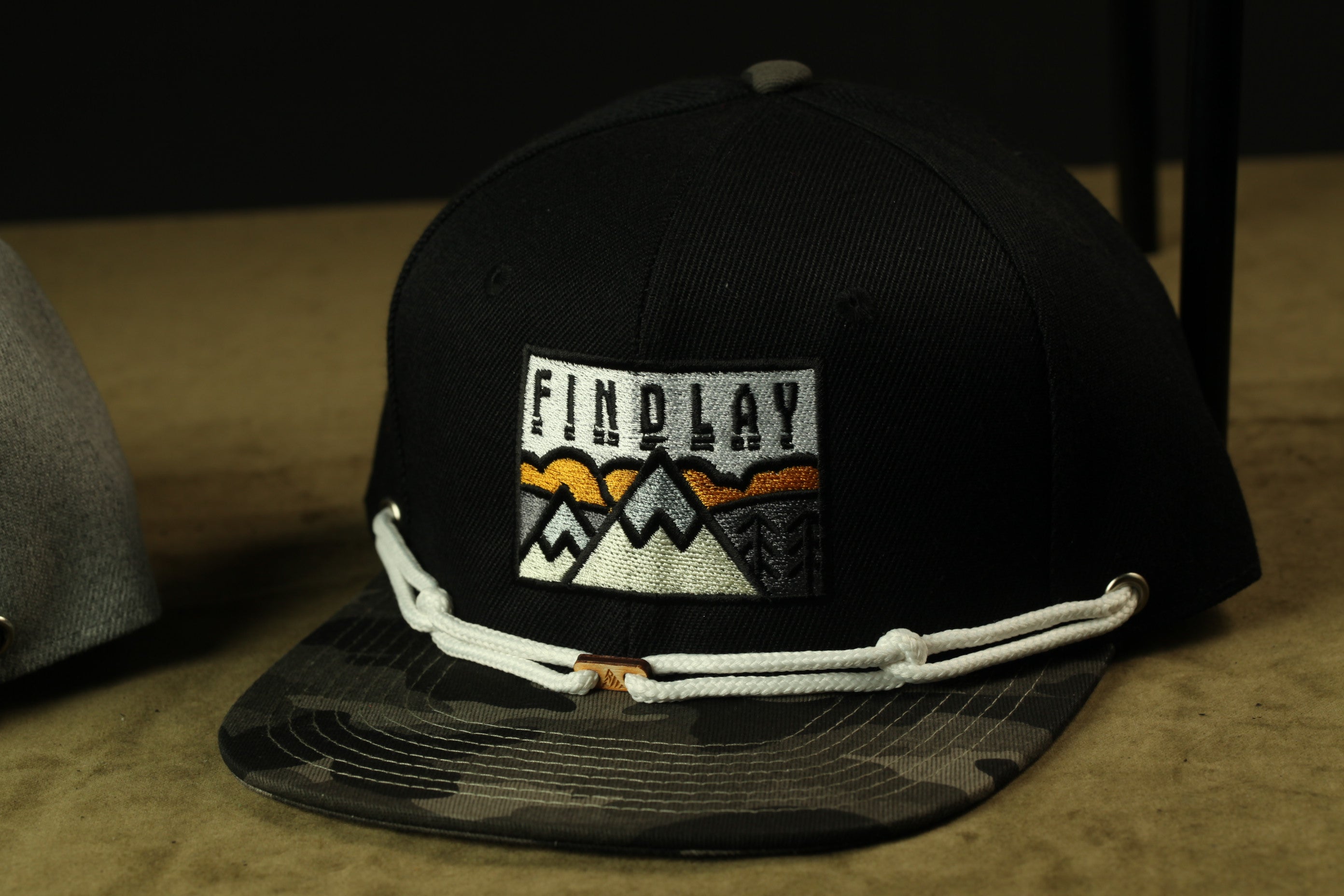 A black baseball cap with a camouflage pattern and a white string around the brim.