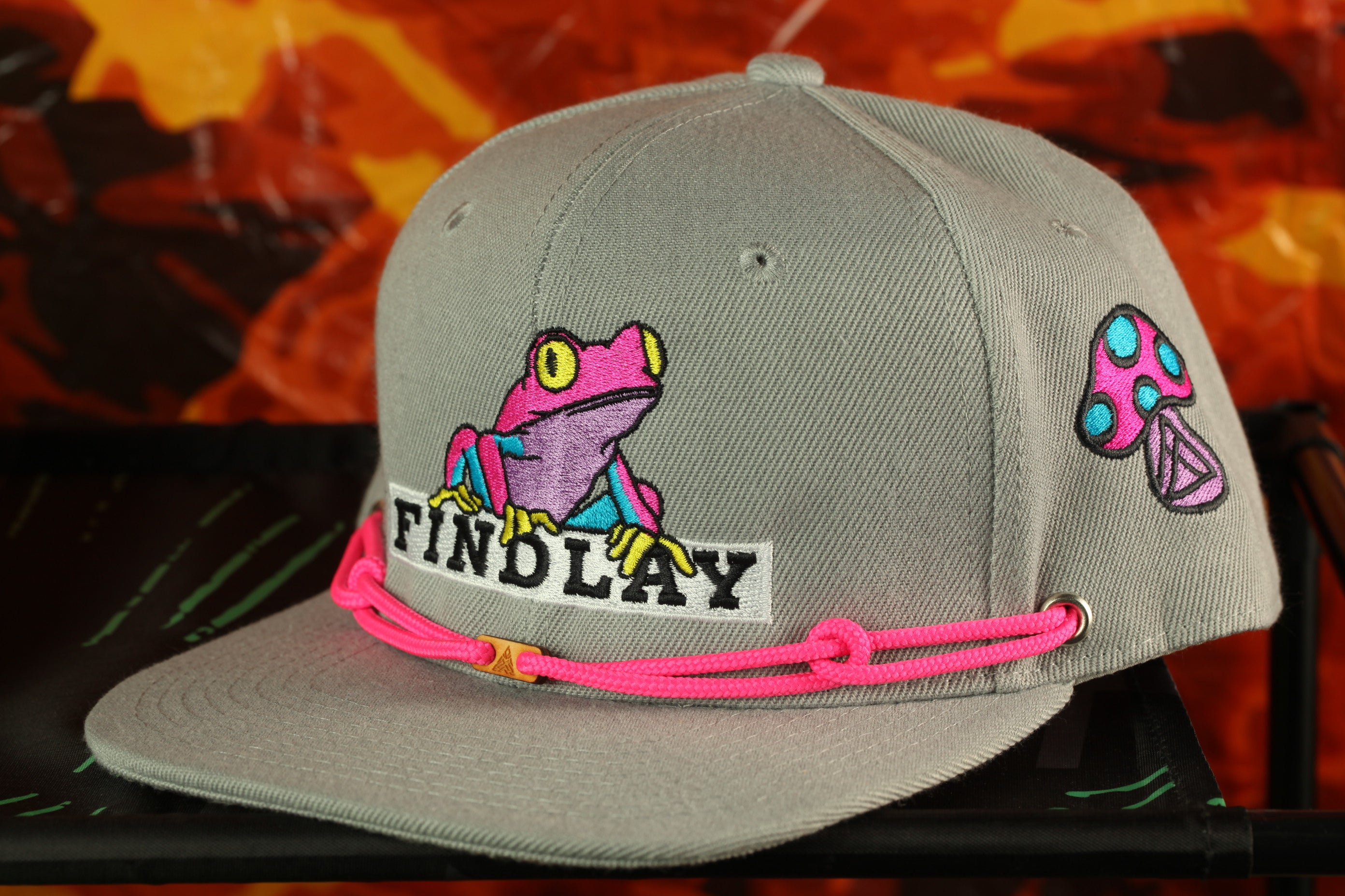 A gray baseball cap with a frog design and the word "FINDLAY" printed on it, along with a pink string or strap.