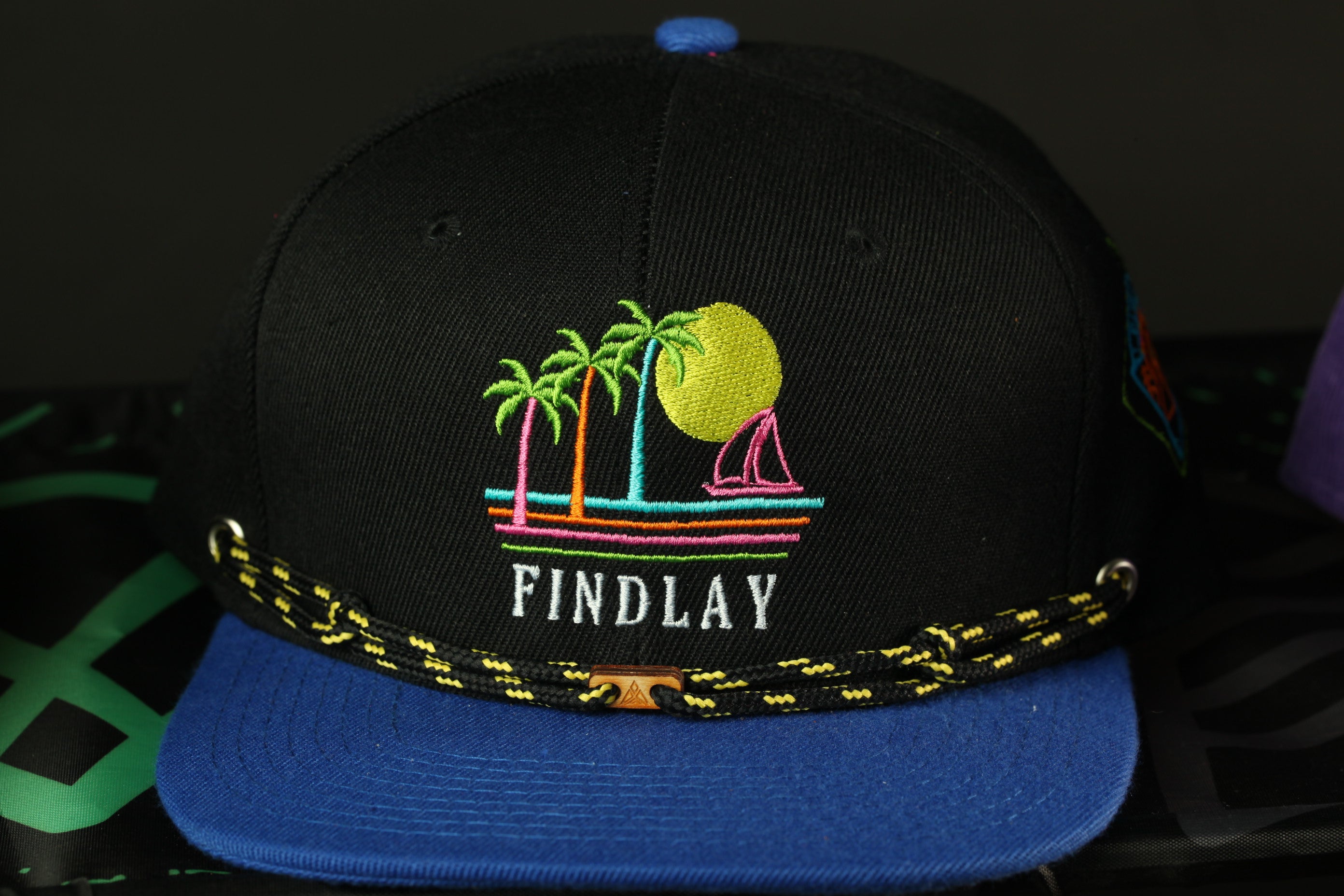 A black baseball cap with a colorful tropical beach scene embroidered on it, featuring palm trees, a yellow sun, and a sailboat.