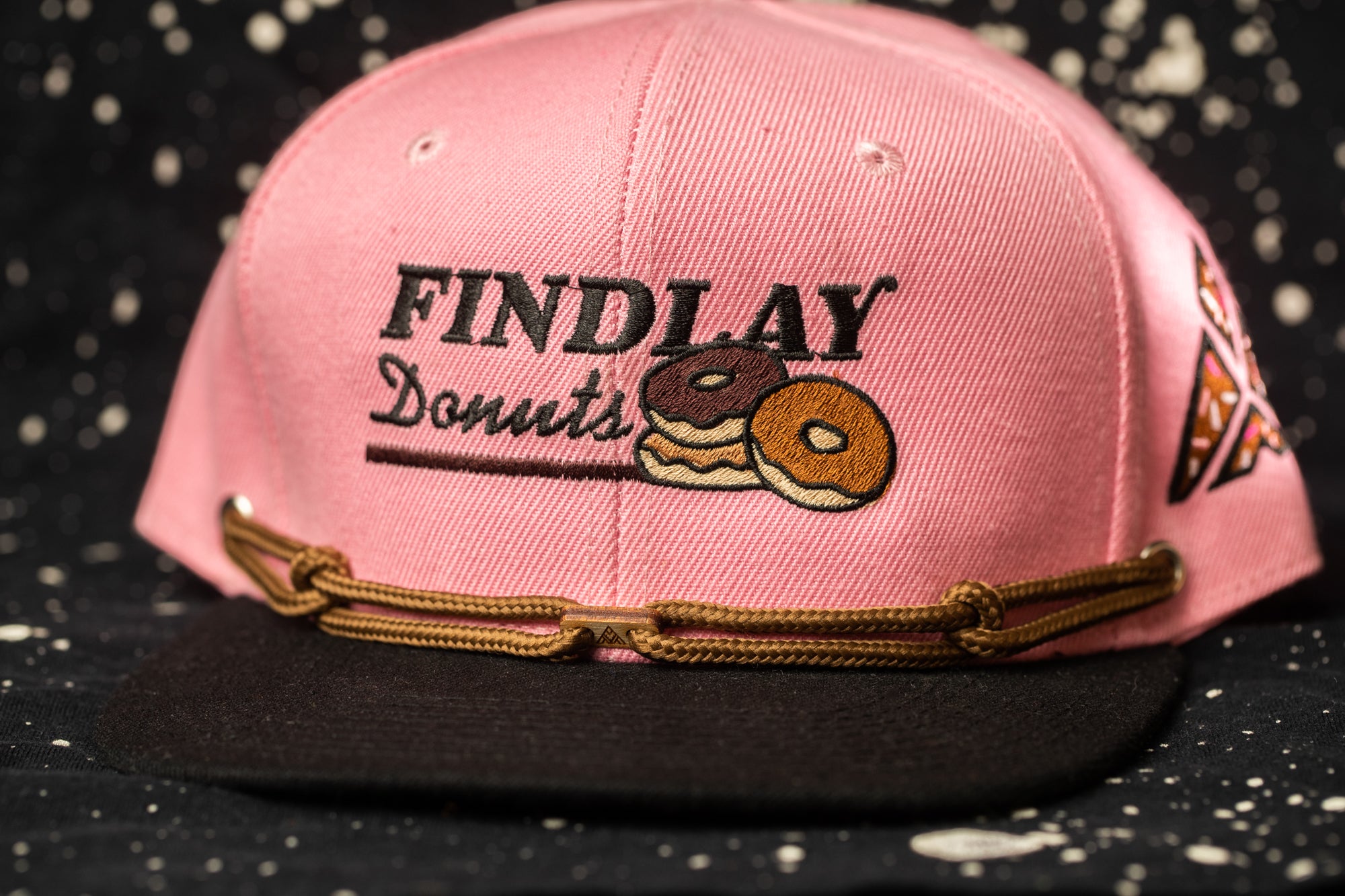A pink baseball cap with the text "Findlay Donuts" printed on it, along with an illustration of two donuts.
