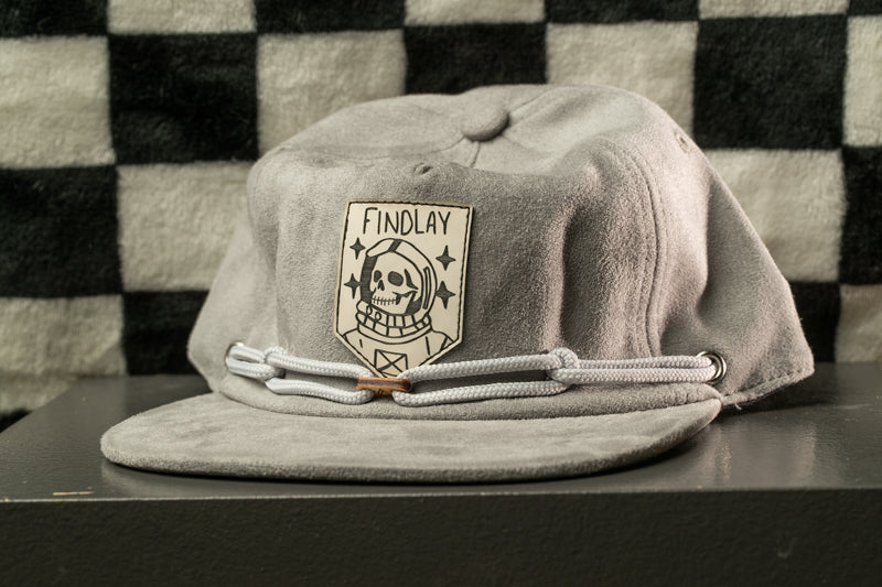 A gray baseball cap with a skull and crossbones design and the text "FINDLAY" printed on it, placed on a black surface against a checkered background.