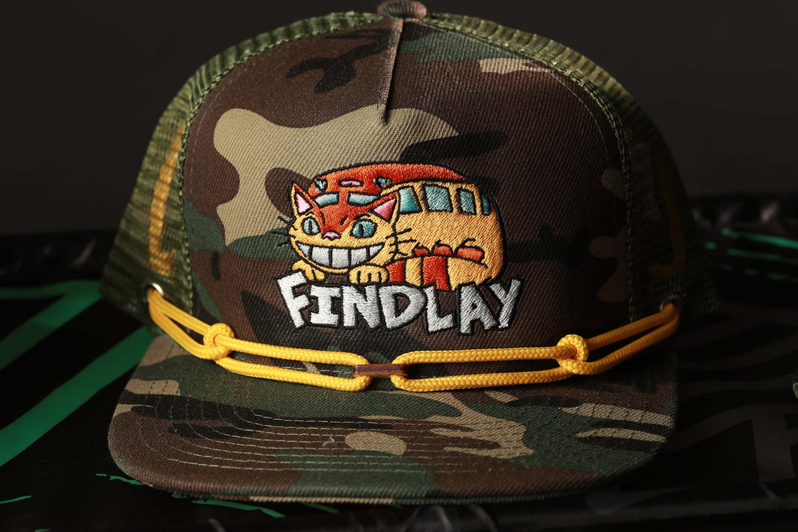 A camouflage baseball cap with a cartoon cat design and the word "Findlay" printed on it.