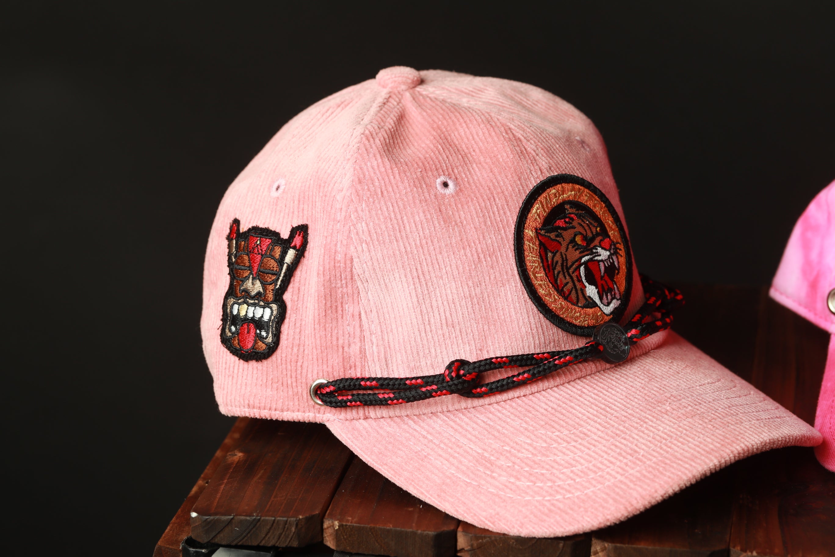 A pink baseball cap with a black and red braided strap, featuring a patch with a skull and crossbones design on the front.
