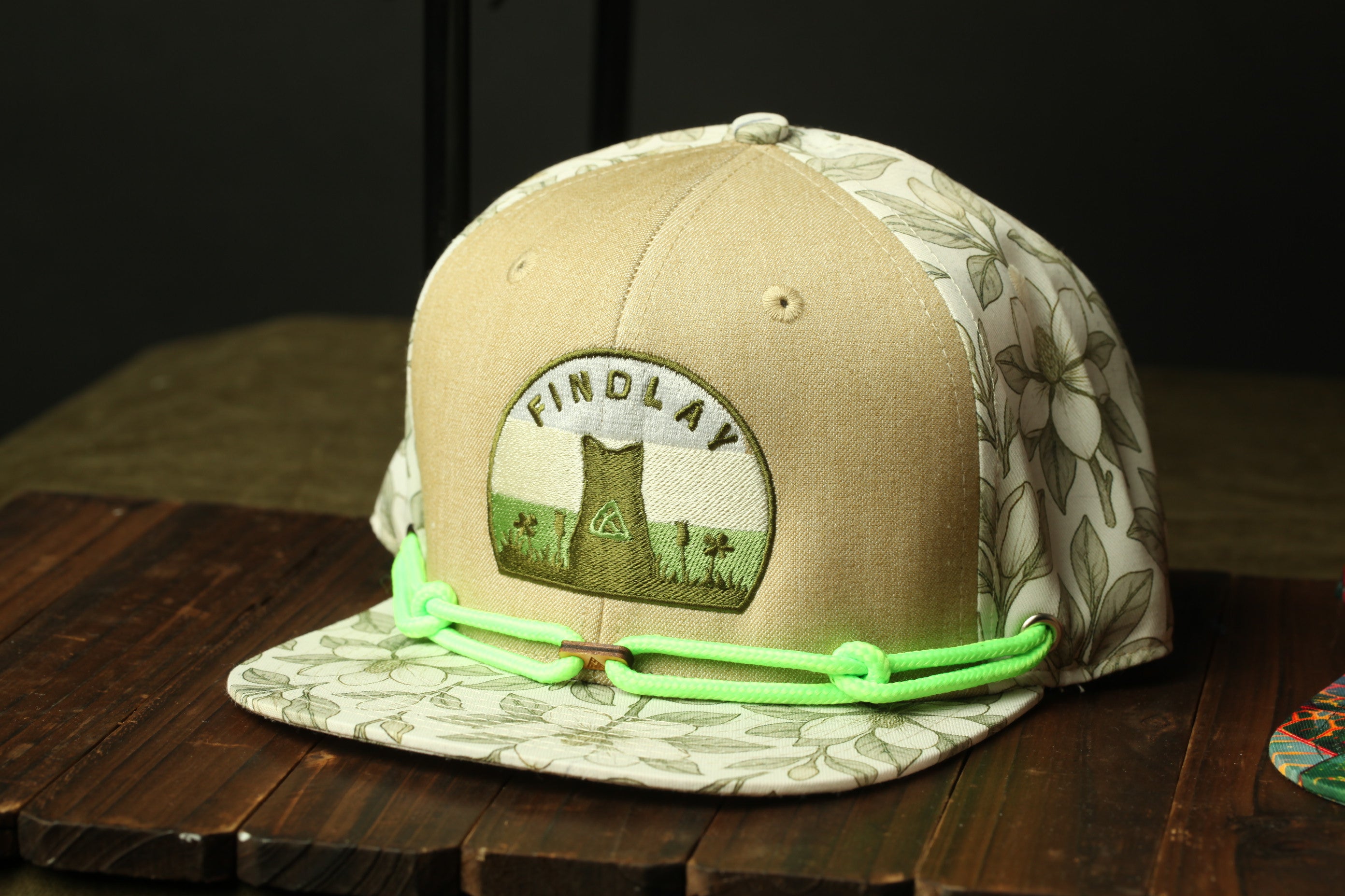 A beige baseball cap with a floral pattern and a patch on the front, sitting on a wooden surface.