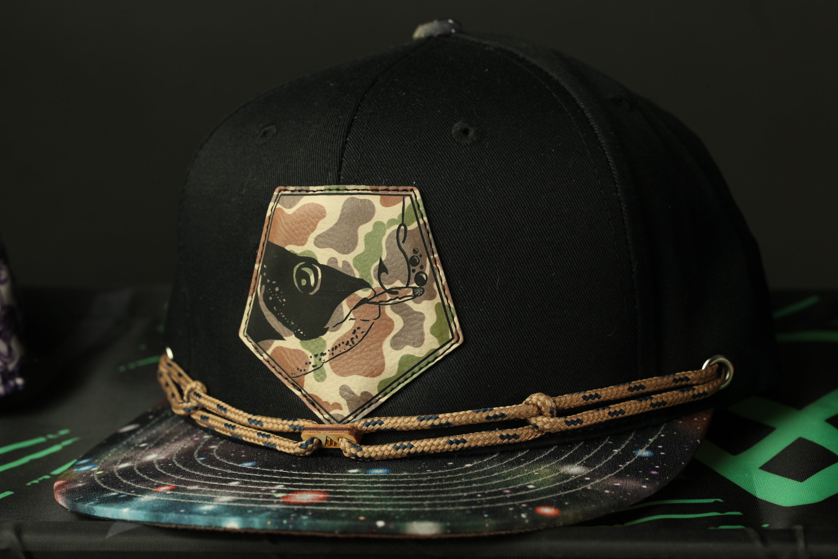 A black baseball cap with a camouflage patch and a space-themed design on the brim, resting on a black surface with a green and black patterned background.