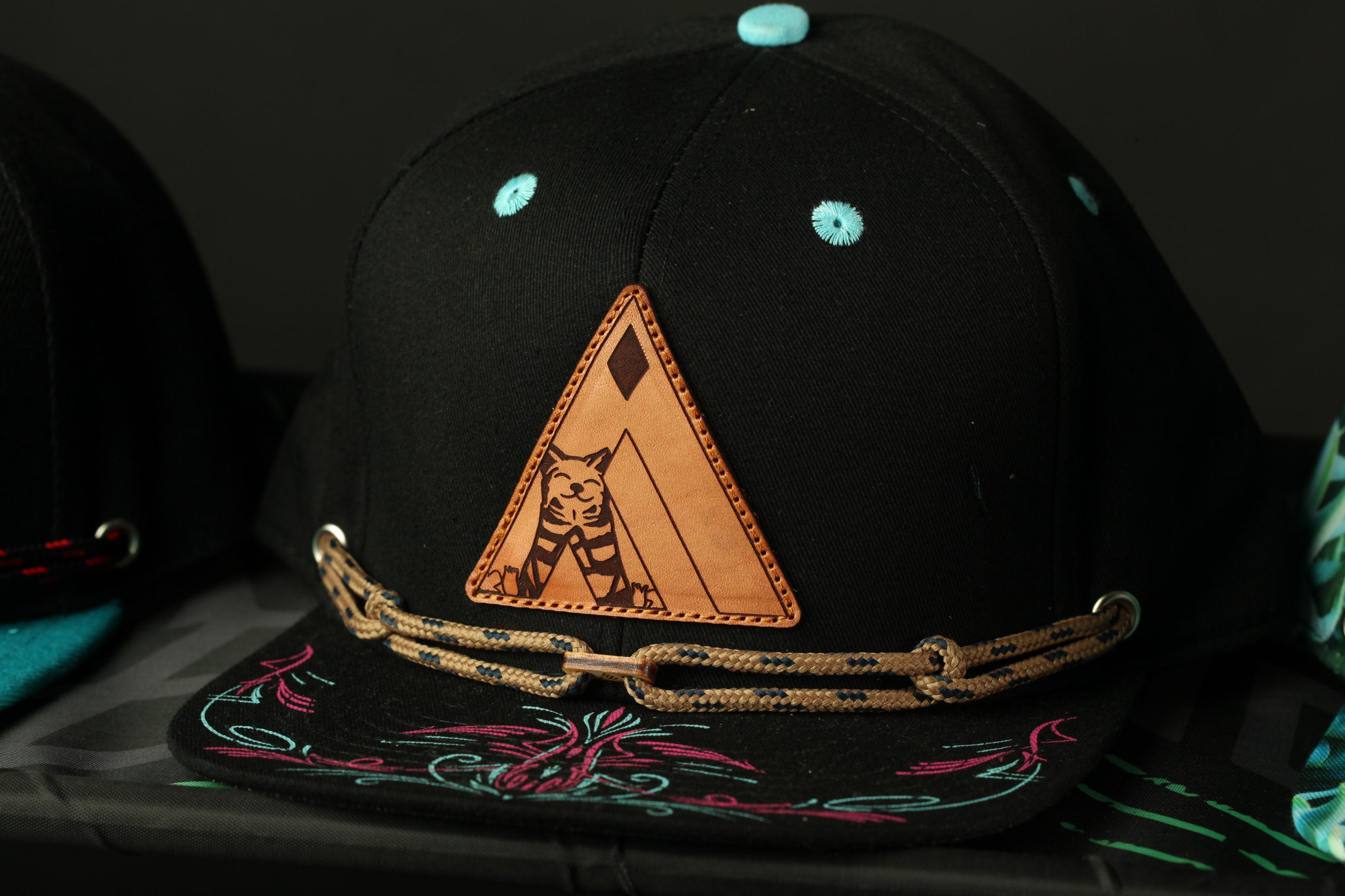A black baseball cap with a triangular patch featuring a cat design, a chain, and a floral pattern.