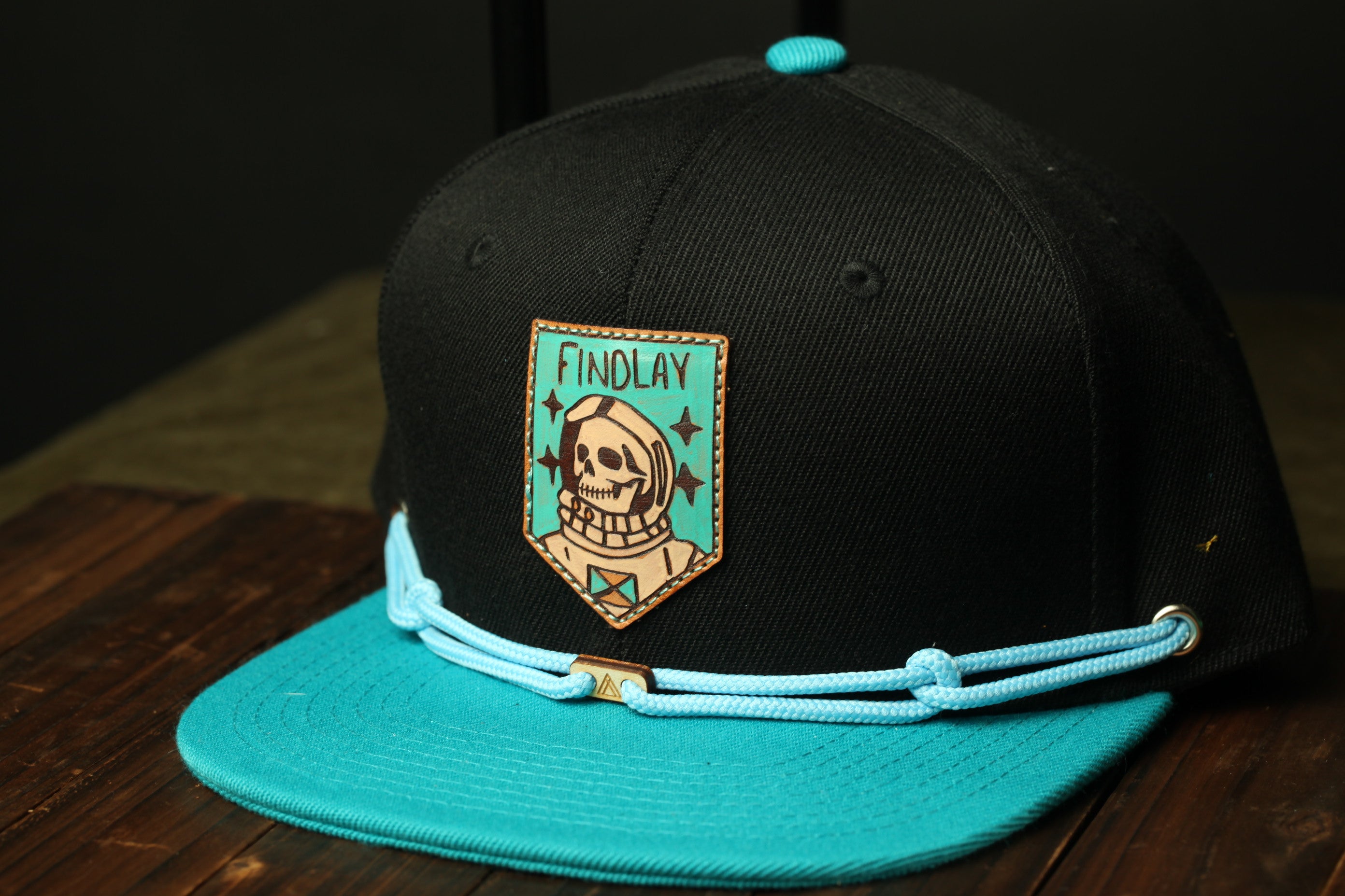 A black baseball cap with a teal brim and a patch featuring a skull and crossbones design.