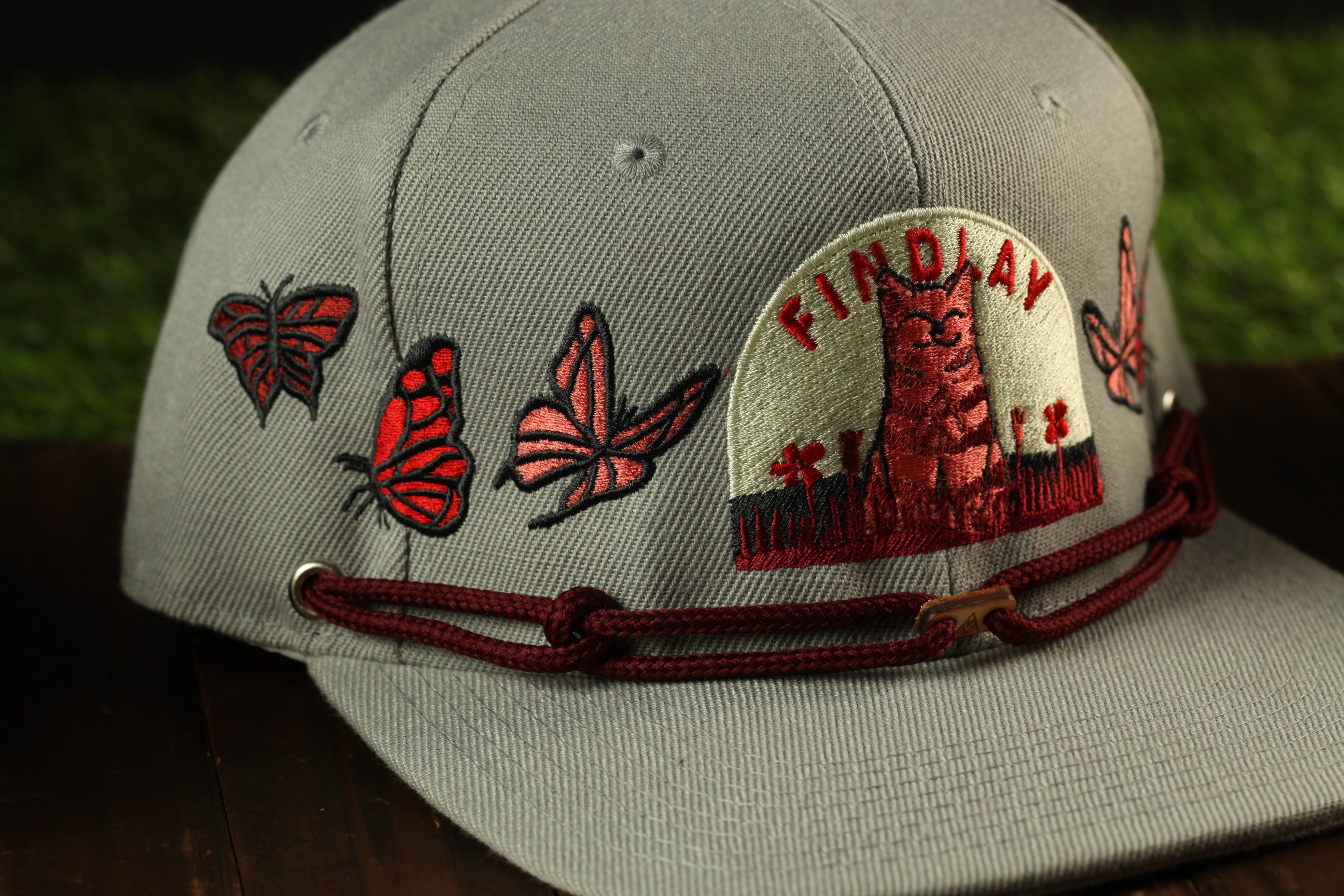 A gray baseball cap with a red and white embroidered design, including a cat and butterflies, and the text "FIND LAY" on the front.