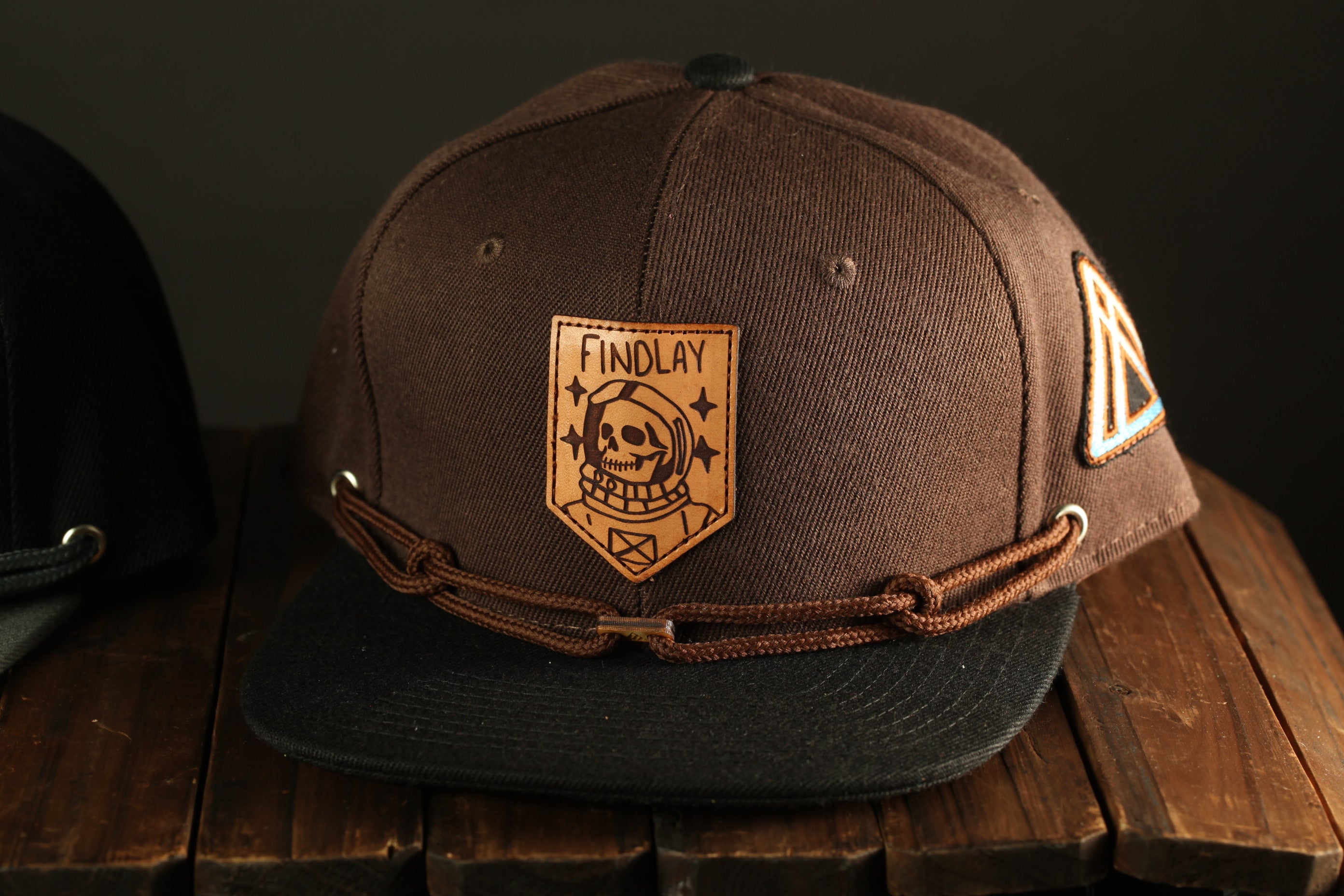 A brown baseball cap with a skull and crossbones design and the word "FINDOLAY" printed on it, sitting on a wooden surface.