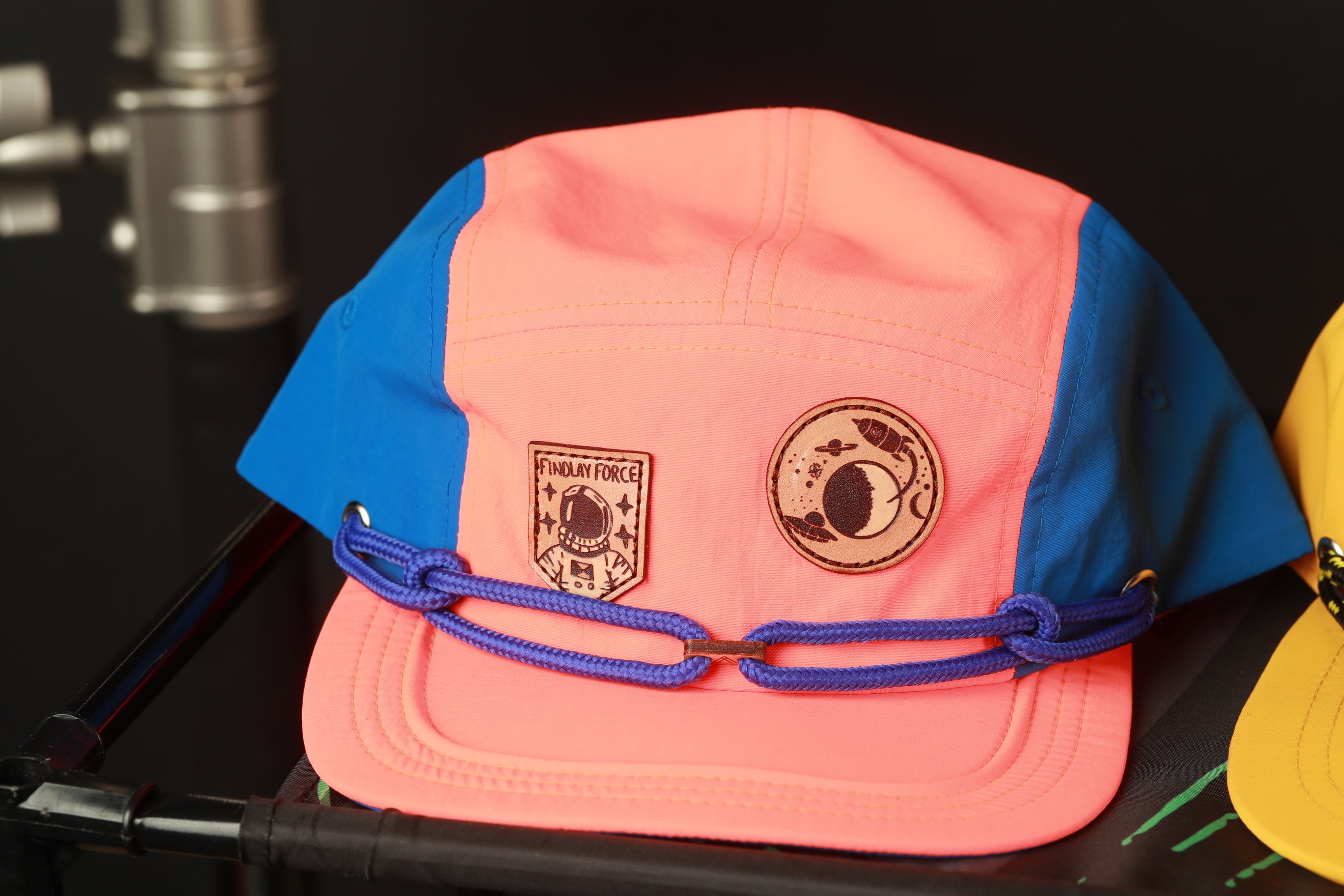 A pink and blue baseball cap with a blue band and a circular patch on the front, sitting on a black surface.