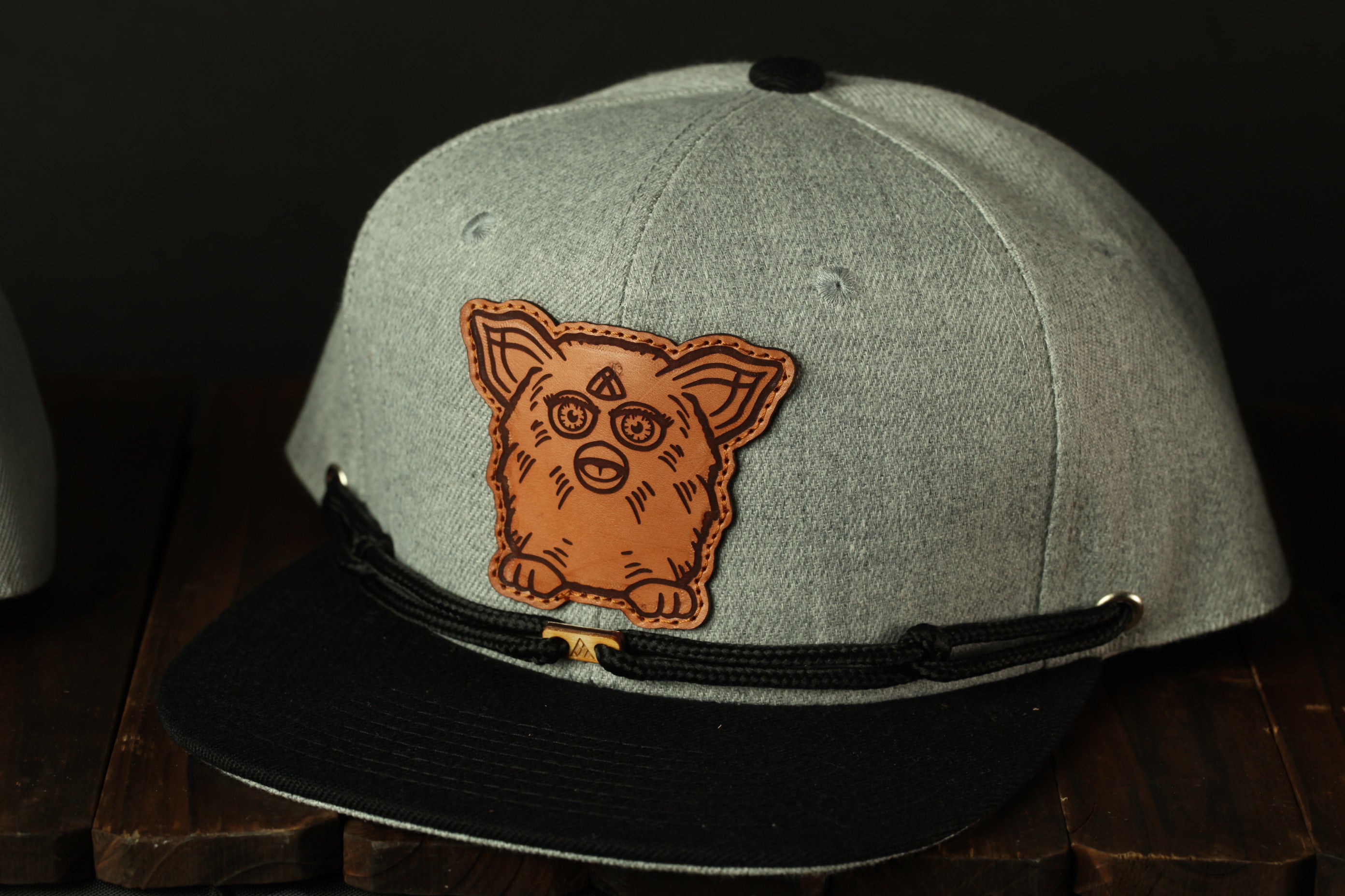 A gray baseball cap with a black brim and a brown embroidered patch featuring a cartoon-like creature on the front.