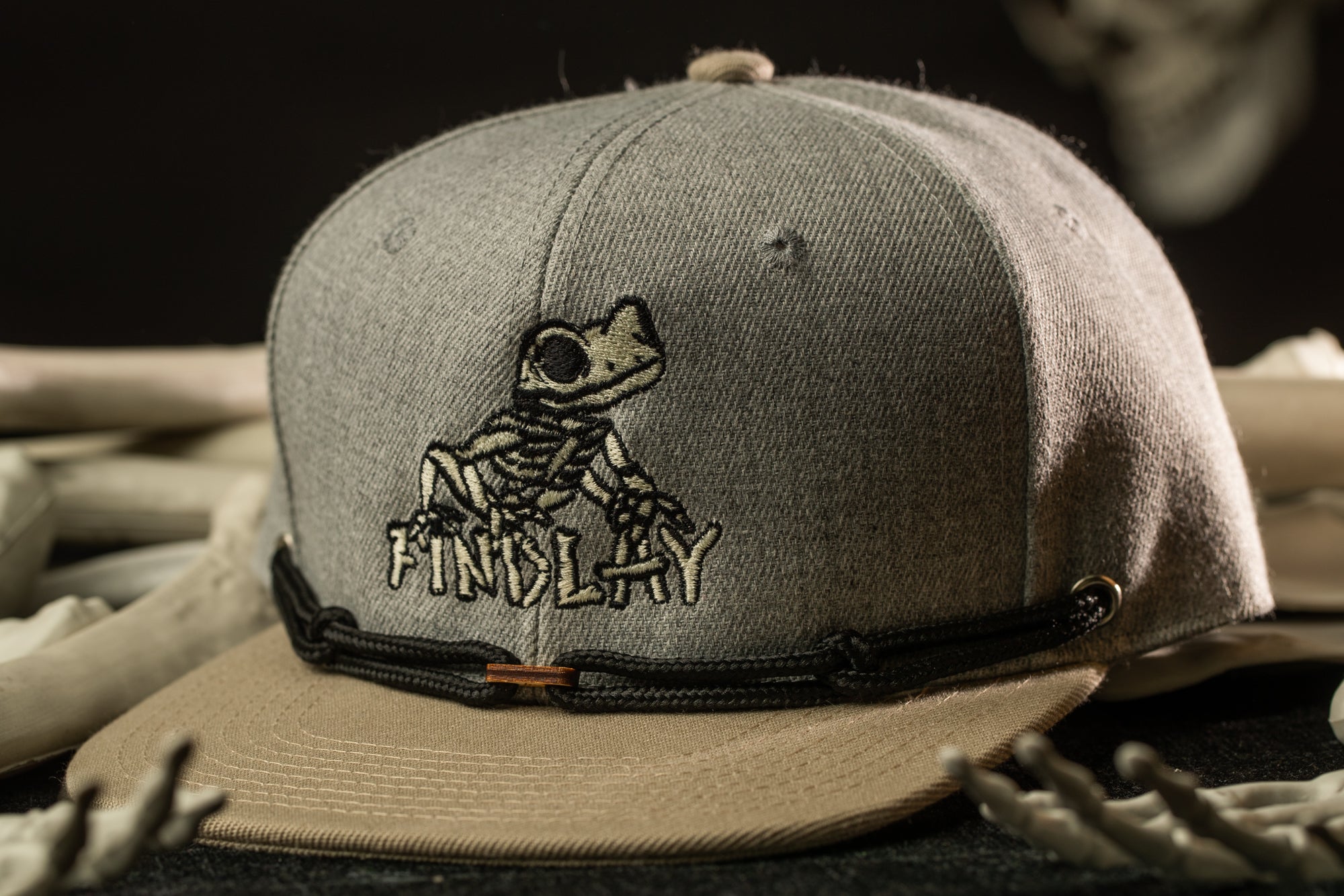 A gray baseball cap with a cartoon frog design and the word "FINSLAY" printed on it, placed on a surface with some bones.
