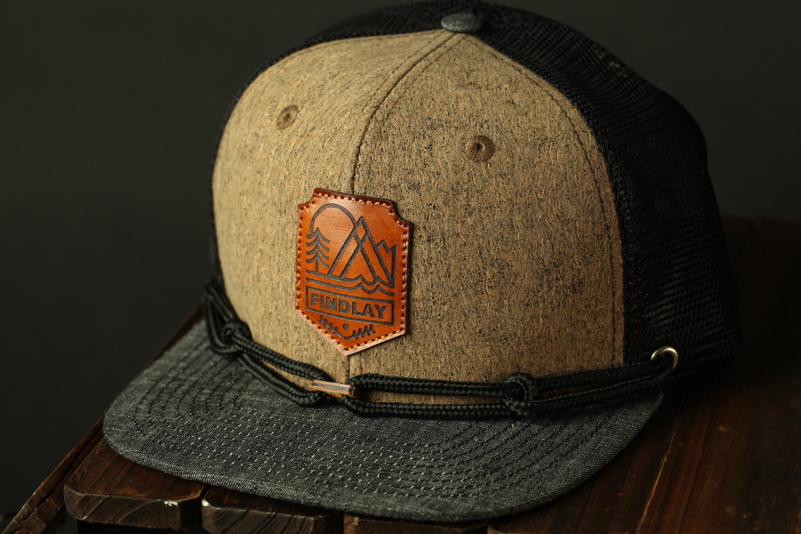 A tan and black baseball cap with a leather patch on the front, featuring a mountain and tree design.
