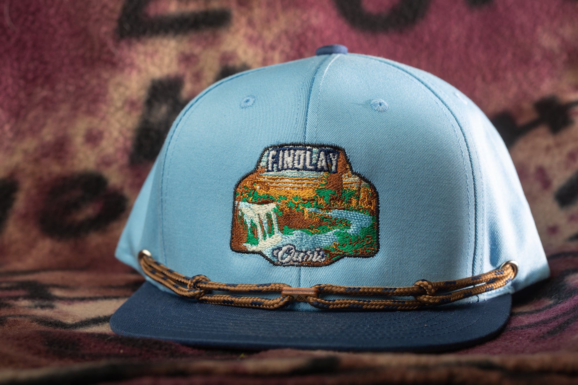 A light blue baseball cap with a decorative patch on the front, featuring a scenic landscape.
