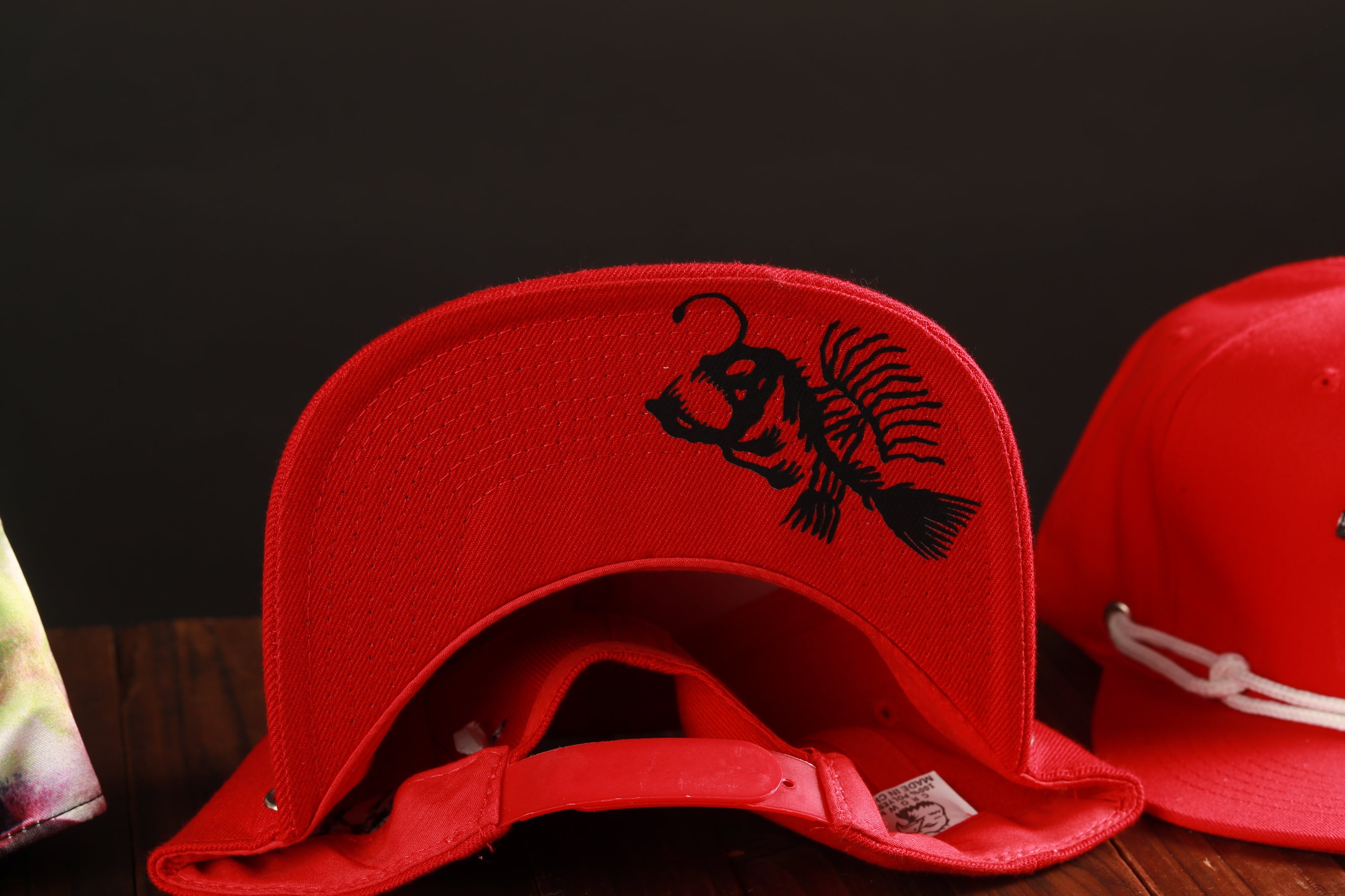 a close-up view of a red baseball cap with a black fish design on it, placed on a wooden surface.