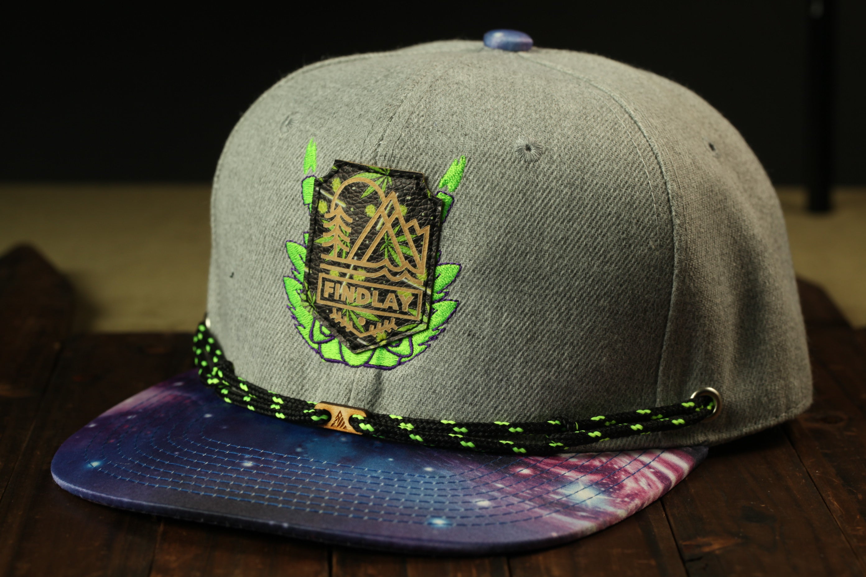 A gray baseball cap with a colorful galaxy print and a green and black braided strap.