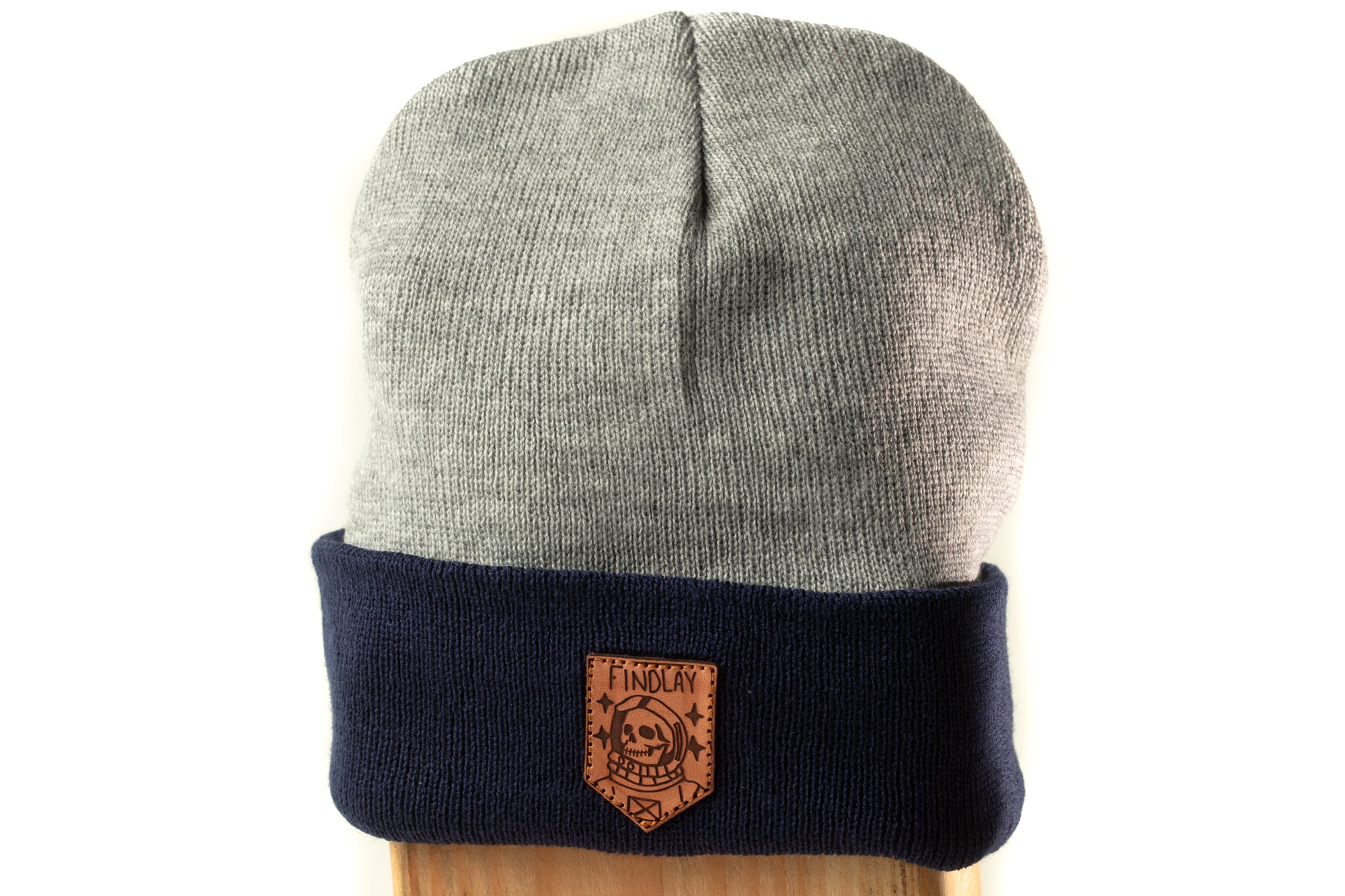 A gray and navy blue beanie with a brown leather patch on the front, featuring a skull and crossbones design.