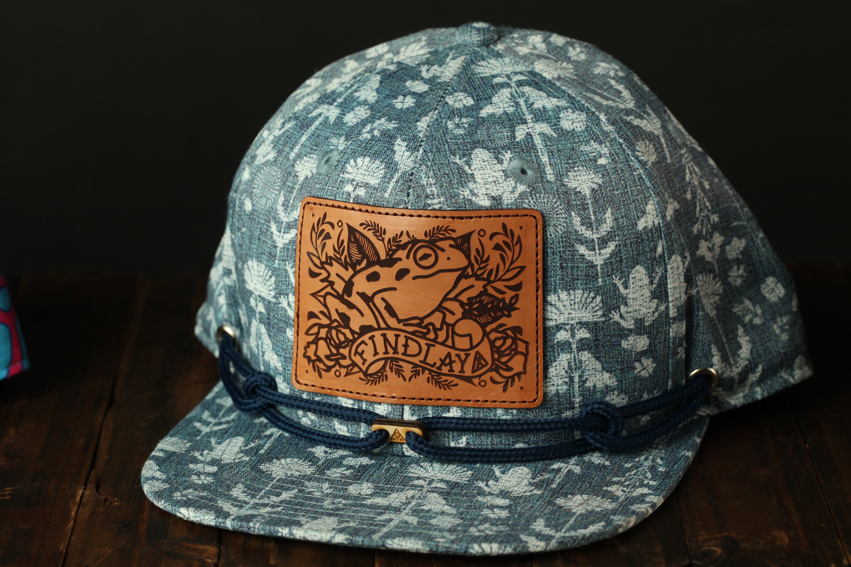 A blue and white patterned baseball cap with a brown leather patch featuring a frog and the text "FIND LAY".