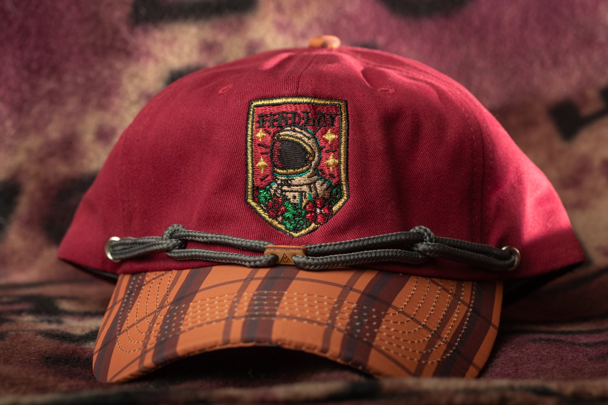 A red baseball cap with a plaid pattern and an embroidered patch on the front, featuring a space-themed design.