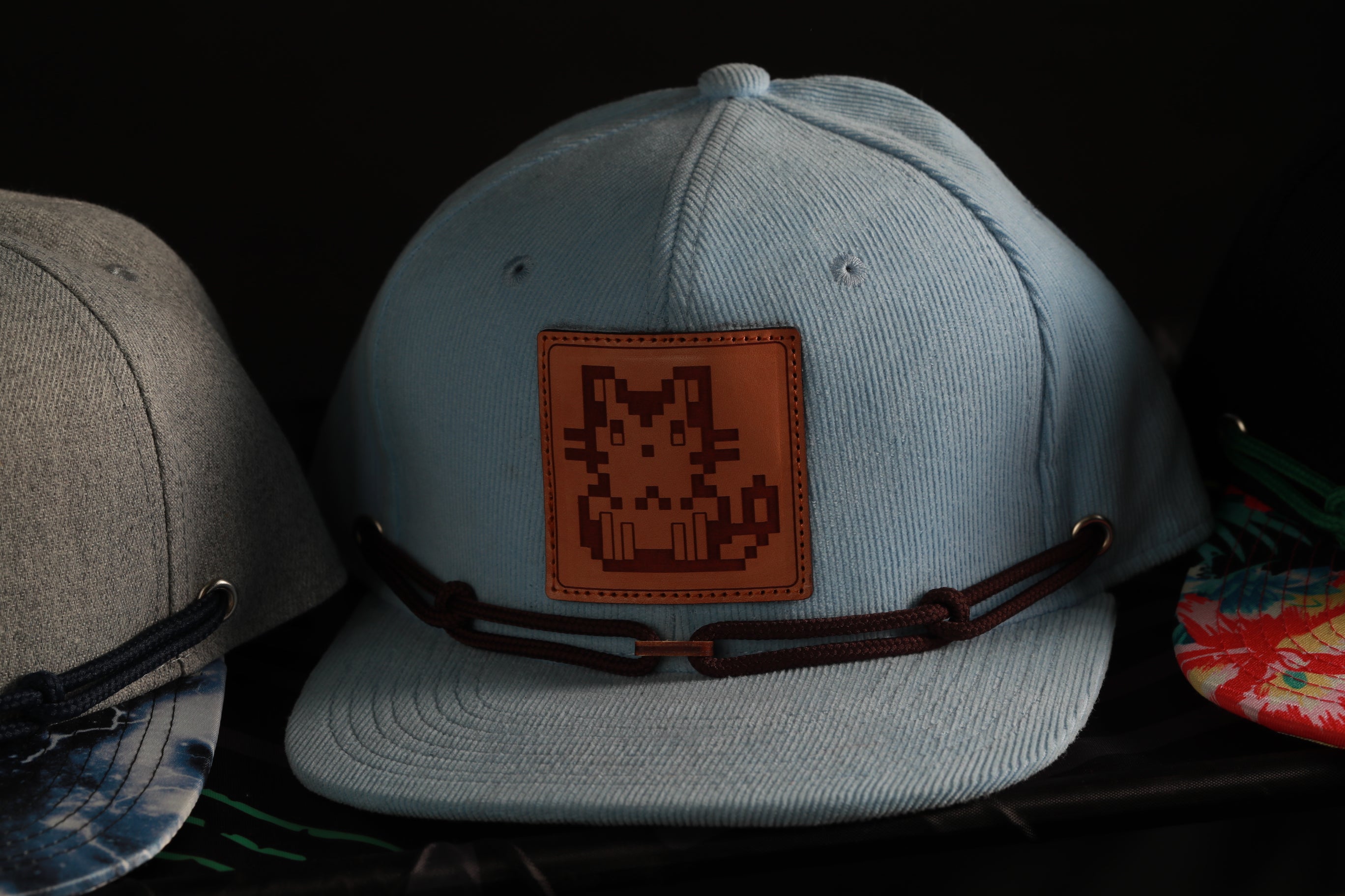 A light blue baseball cap with a cat-like design on the front, featuring a brown leather patch with a pixelated cat image.