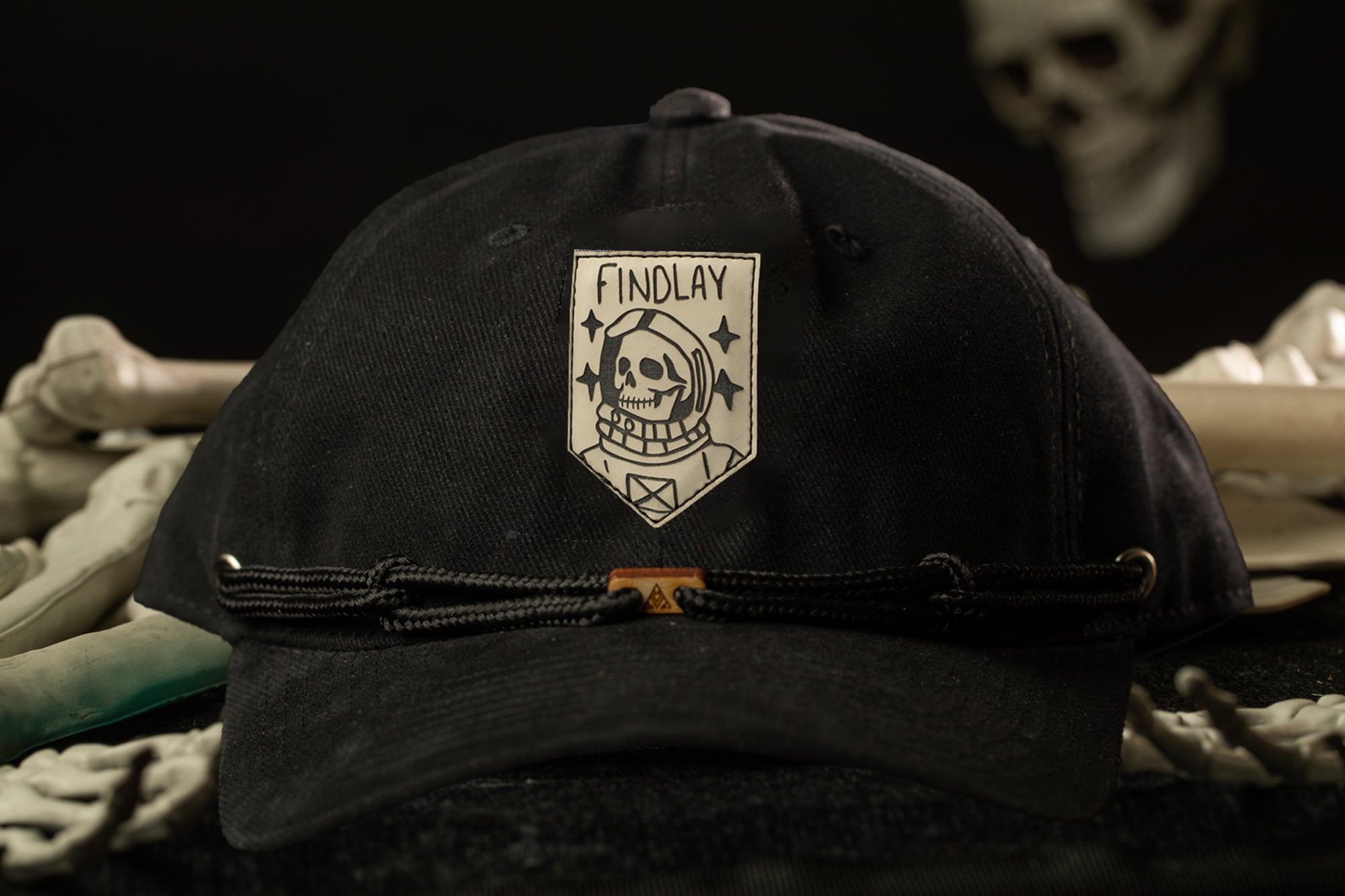 A black baseball cap with a skull and crossbones design and the word "FINDLAY" printed on it, placed on a surface with some bones and a skull visible in the background.