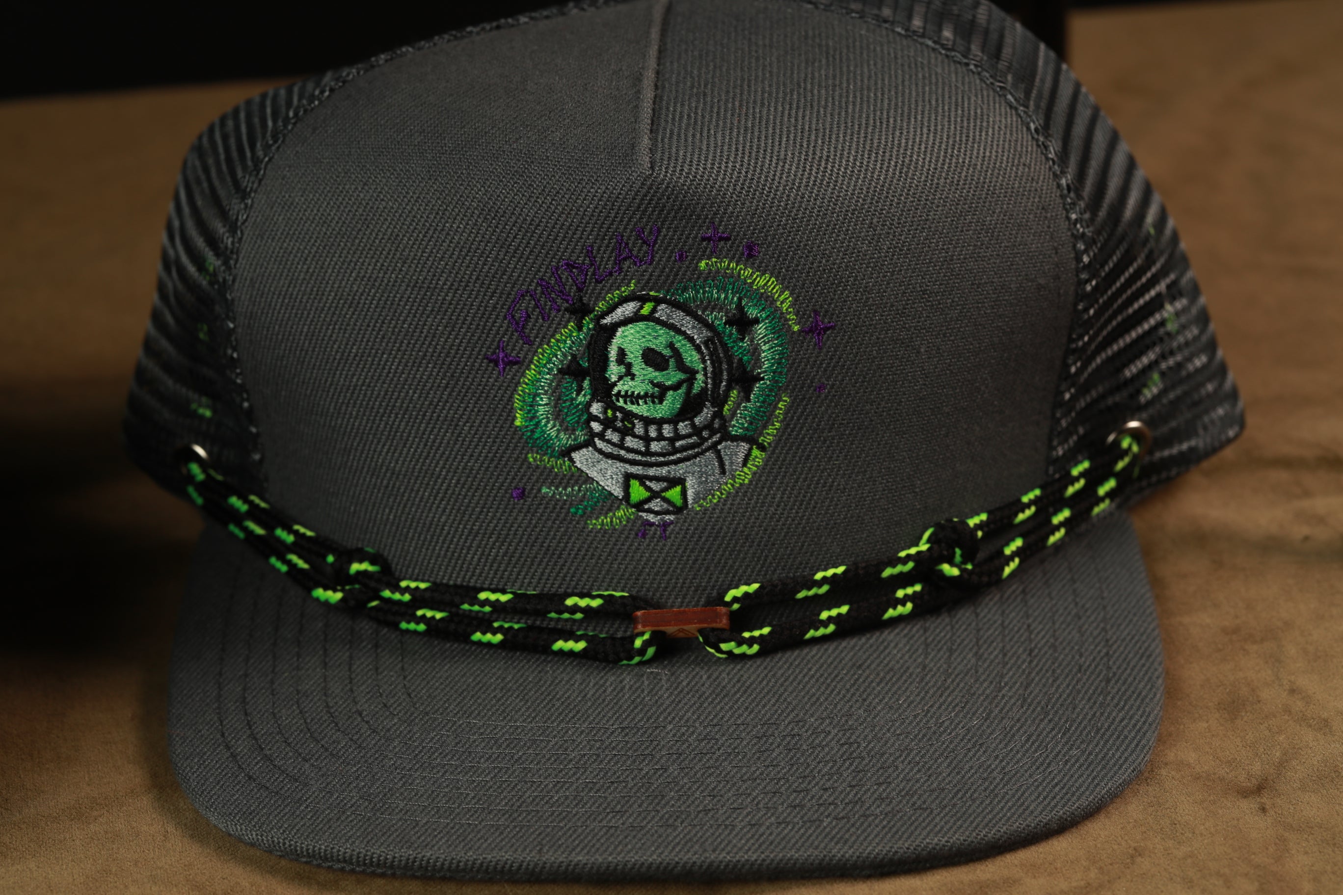 A gray baseball cap with a green and black braided strap, featuring an embroidered design of a skull wearing a spacesuit.