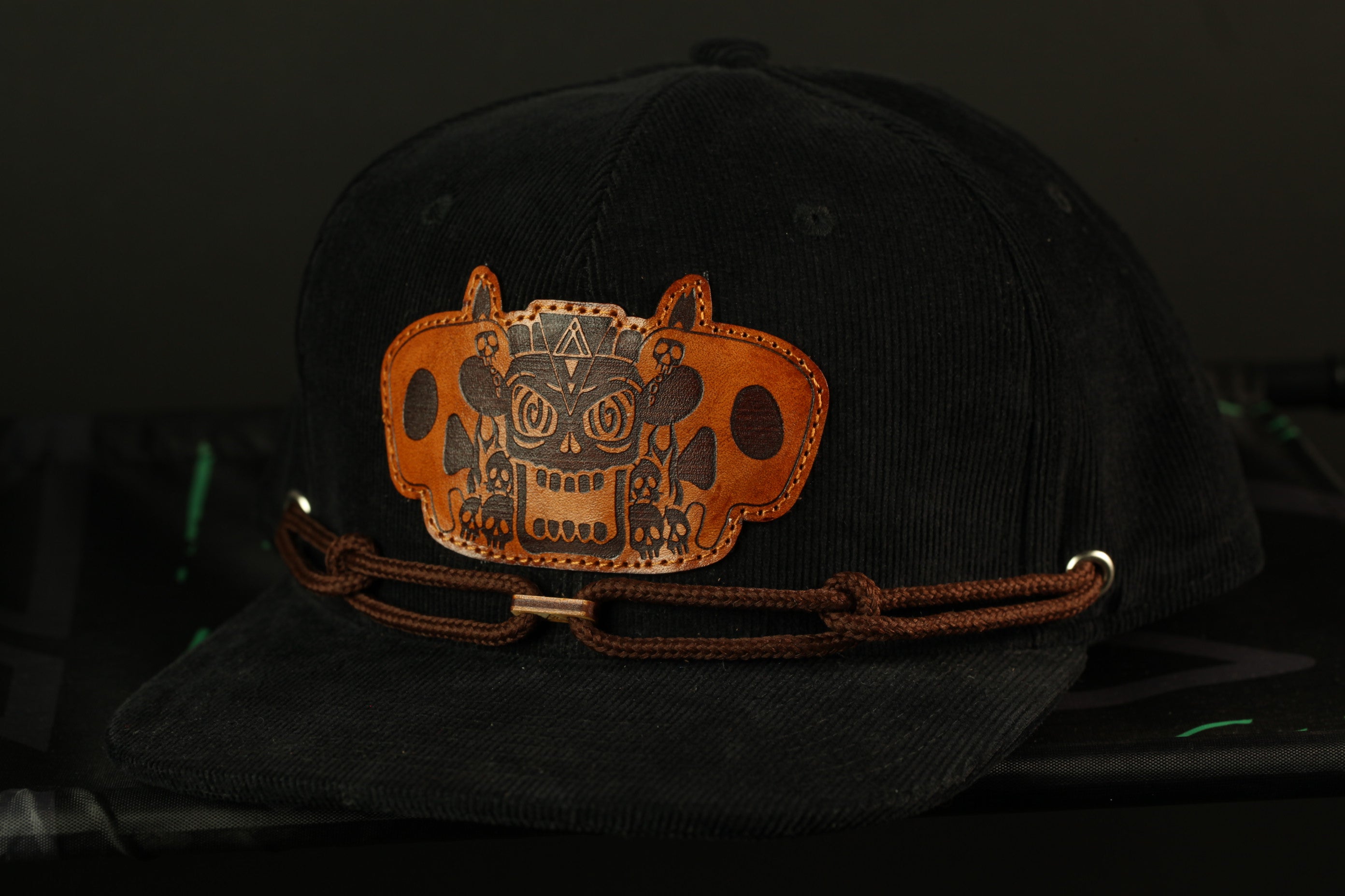 A black baseball cap with a brown leather patch featuring an abstract design on the front.