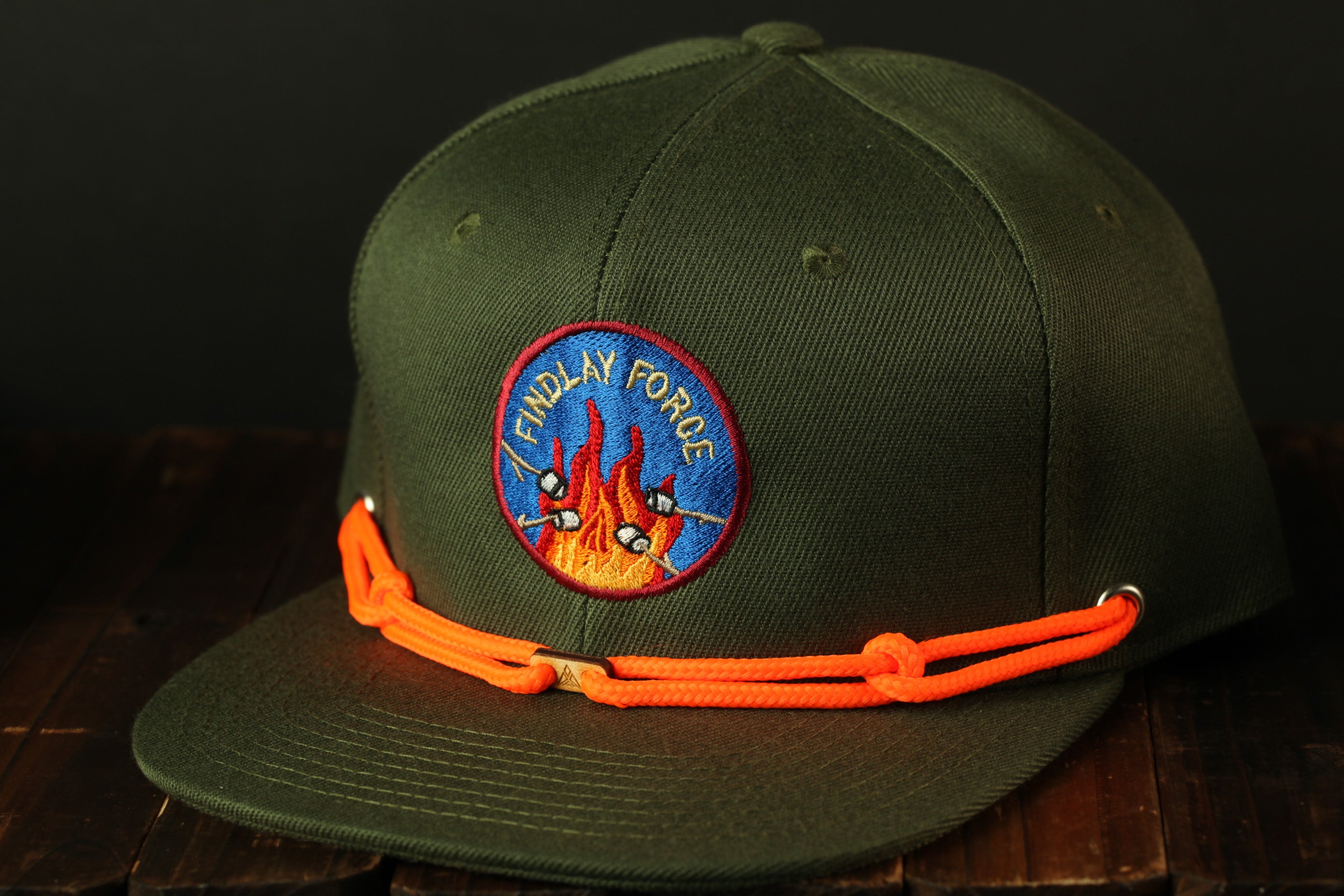 A green baseball cap with an orange string and a circular patch on the front, featuring a flame design.