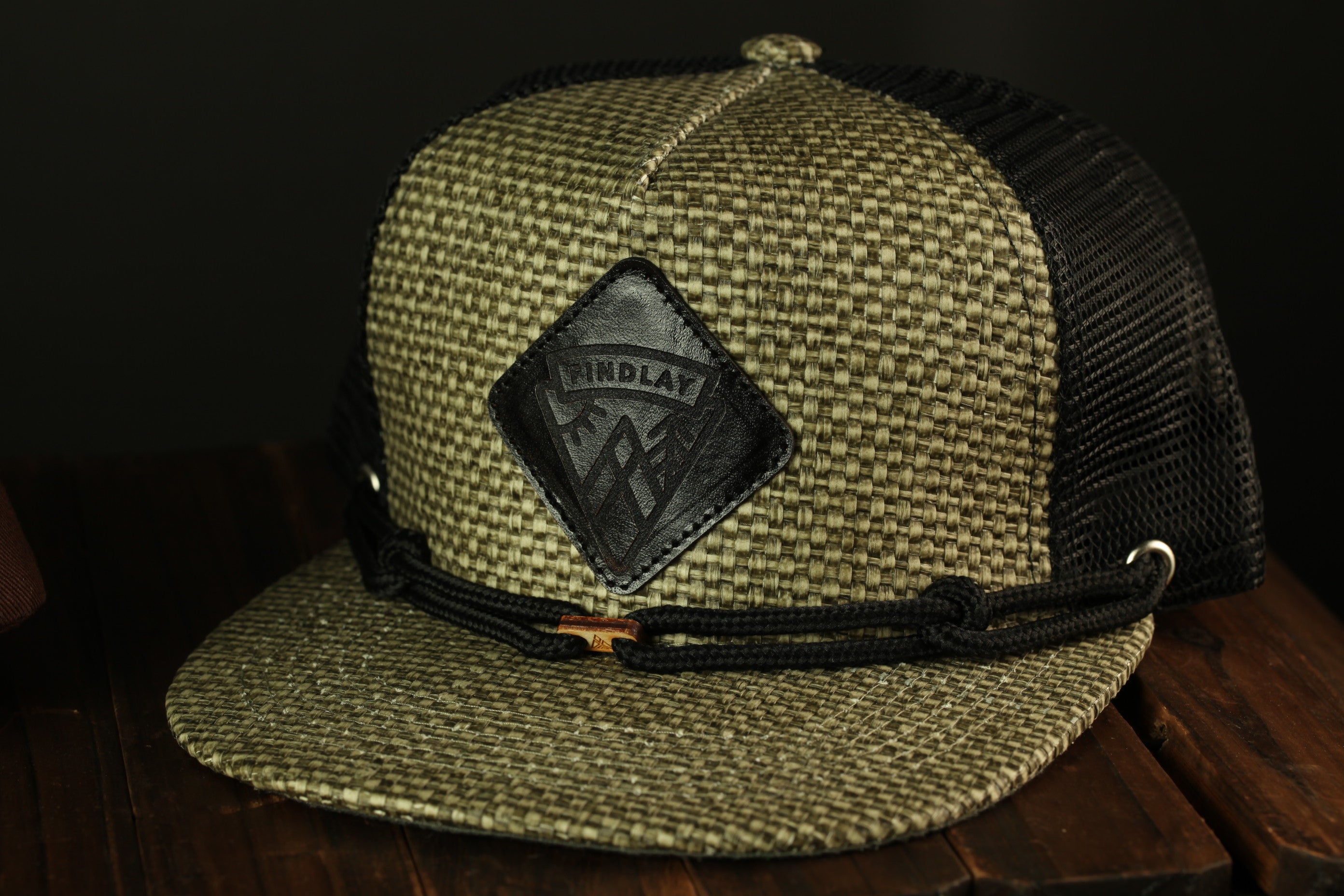 A beige and black woven baseball cap with a black leather patch on the front, sitting on a wooden surface.